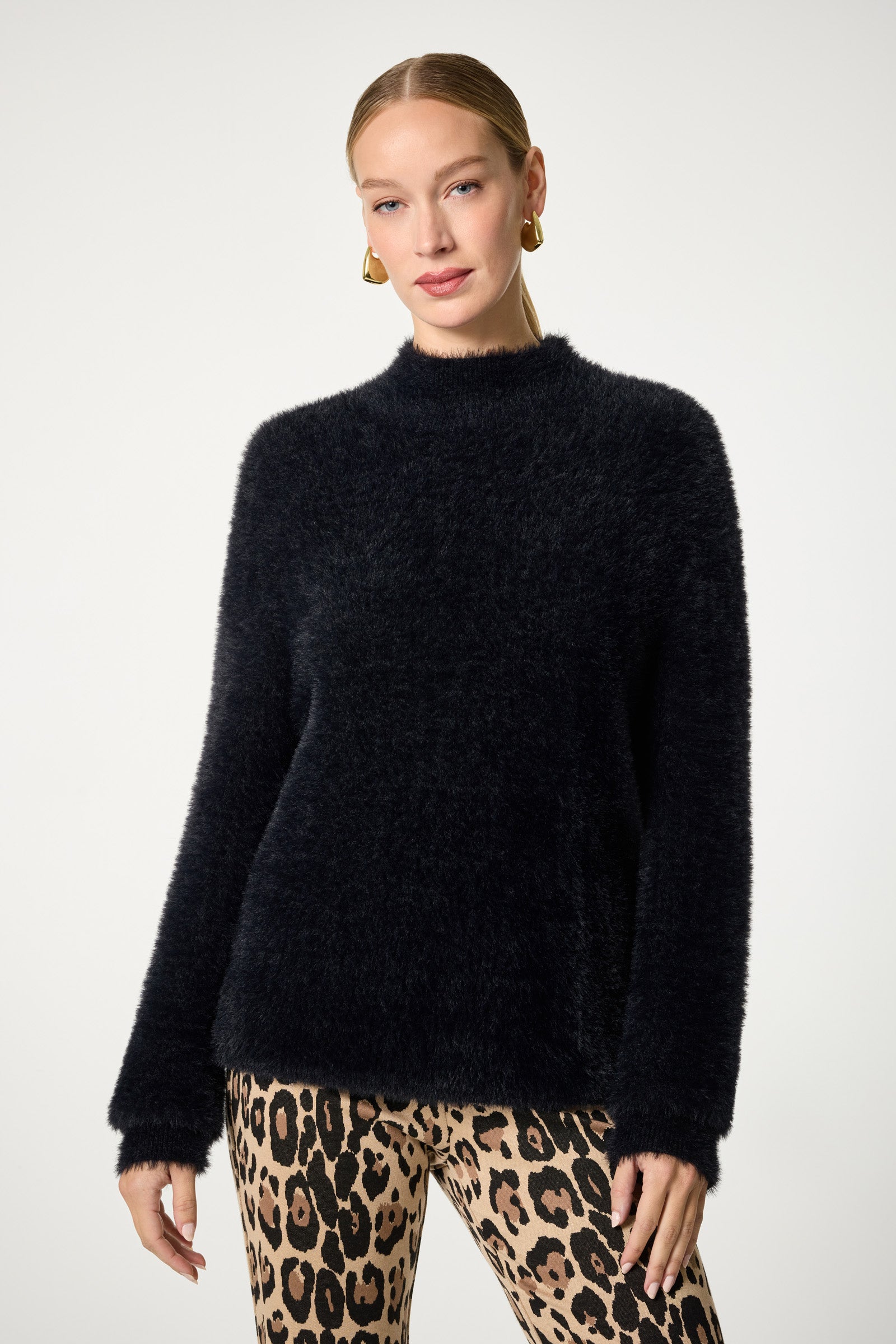 A woman stands against a plain background, wearing the Bazaar Mock Neck Sweater in black with long sleeves and fuzzy texture, paired with leopard print pants. She accessorizes with gold hoop earrings and has her hair pulled back.