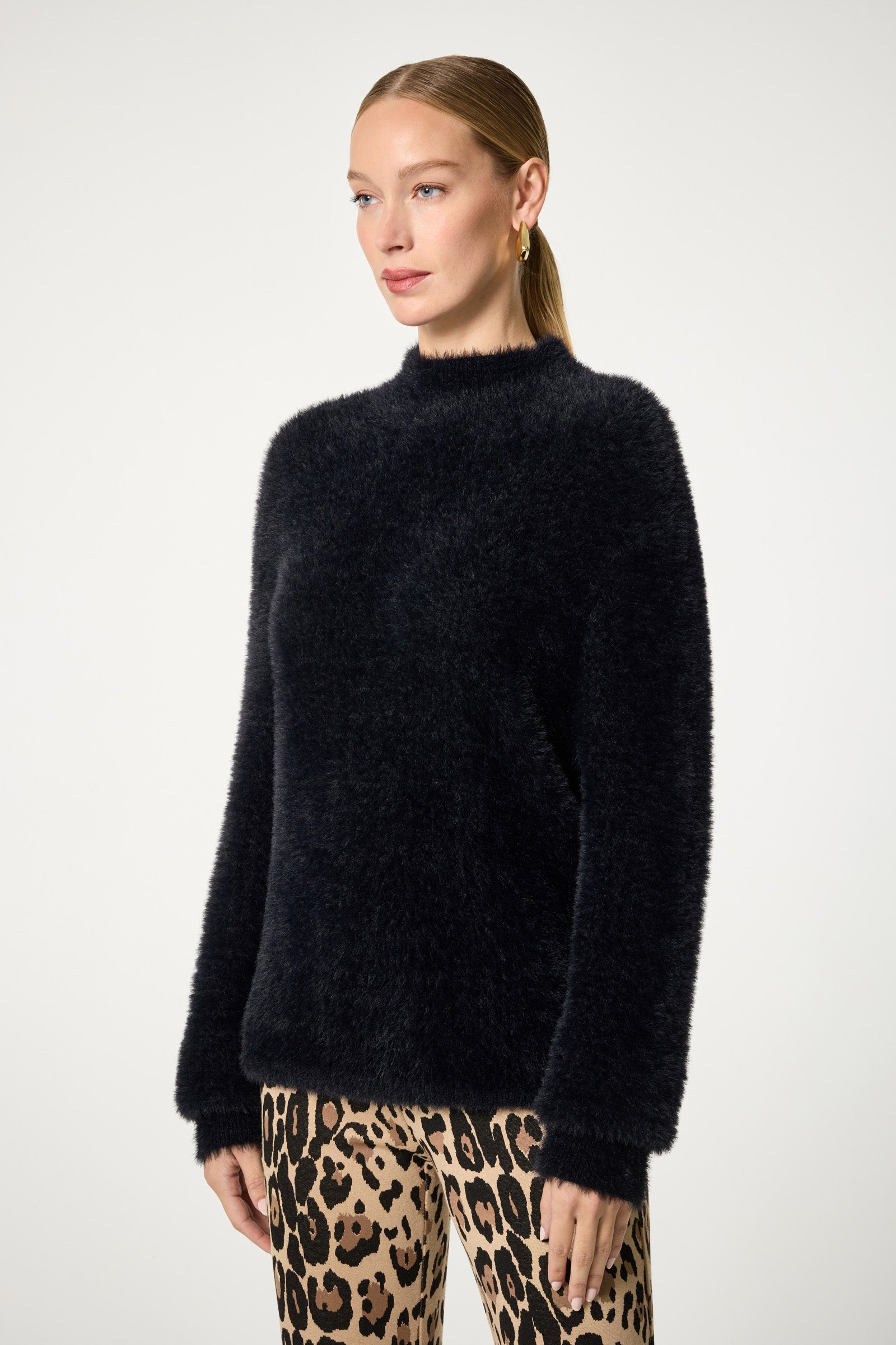 A woman with straight, light brown hair in a low ponytail models the Bazaar Mock Neck Sweater—a fuzzy black long-sleeve style—paired with leopard print pants against a plain light background.