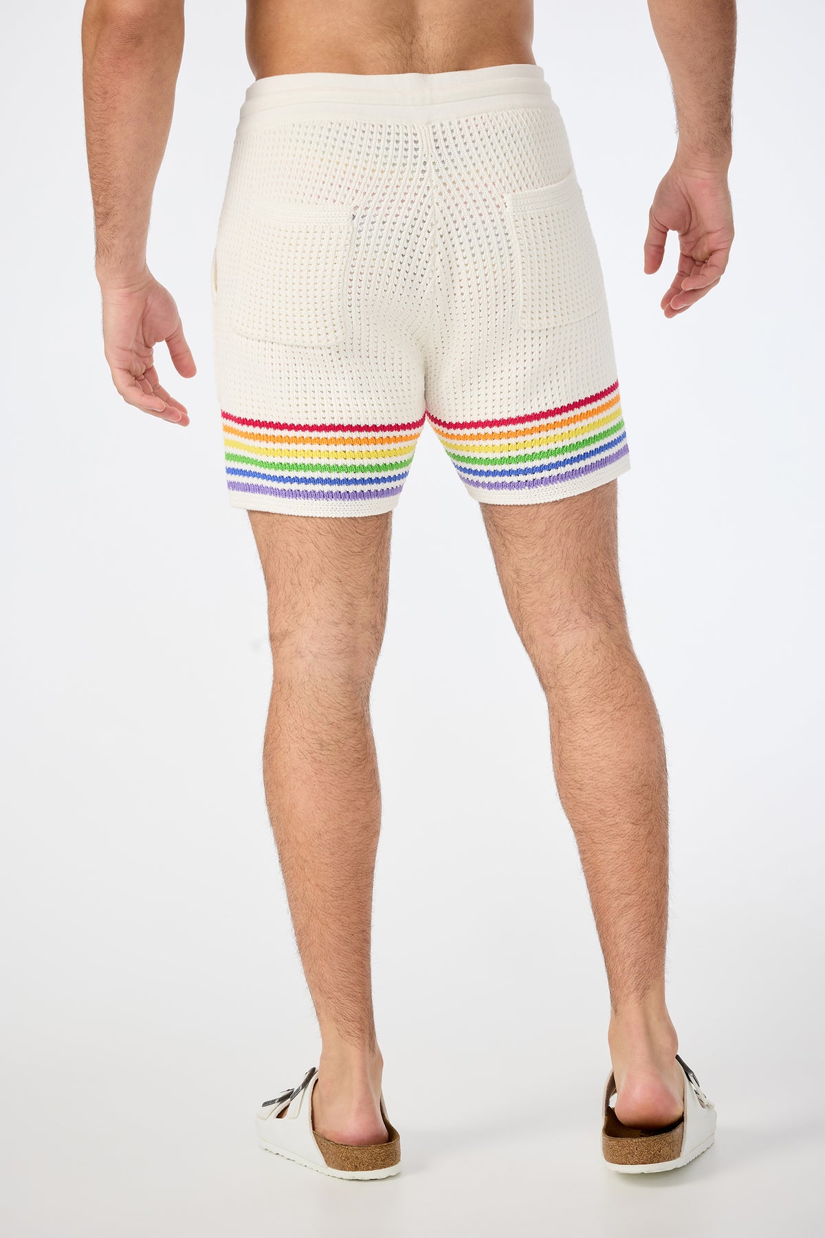 A person stands facing away, wearing the August Open-Knit Short - Pride in cream with rainbow stripes at the hem and white slip-on sandals, set against a plain white background.