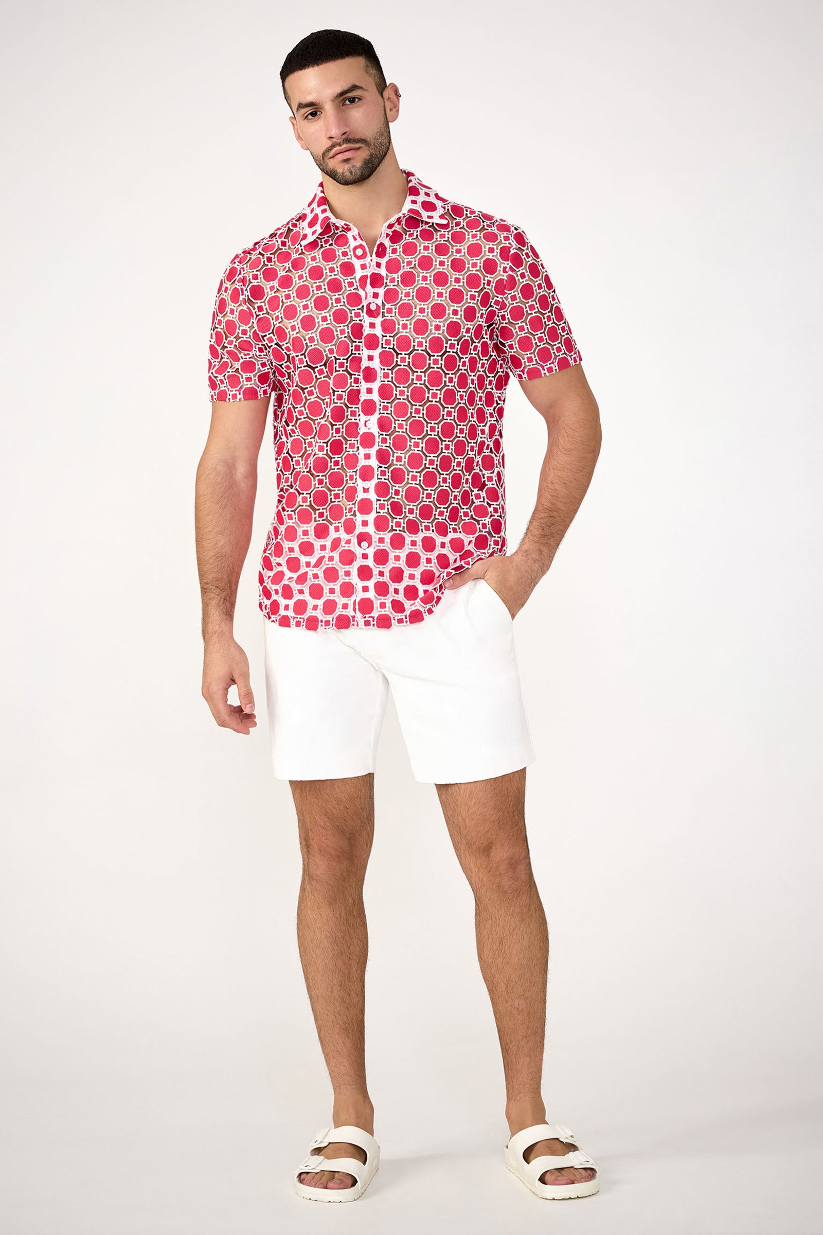 A man stands against a white background wearing the Long Beach Short-Sleeve Shirt in red, paired with white shorts and white sandals. One hand is in his pocket and he has a neutral expression.