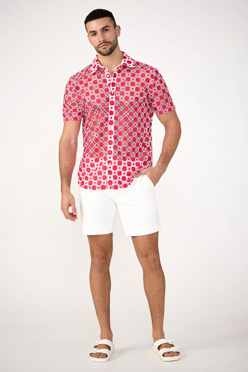 A man stands against a white background wearing the Long Beach Short-Sleeve Shirt in red, paired with white shorts and white sandals. One hand is in his pocket and he has a neutral expression.