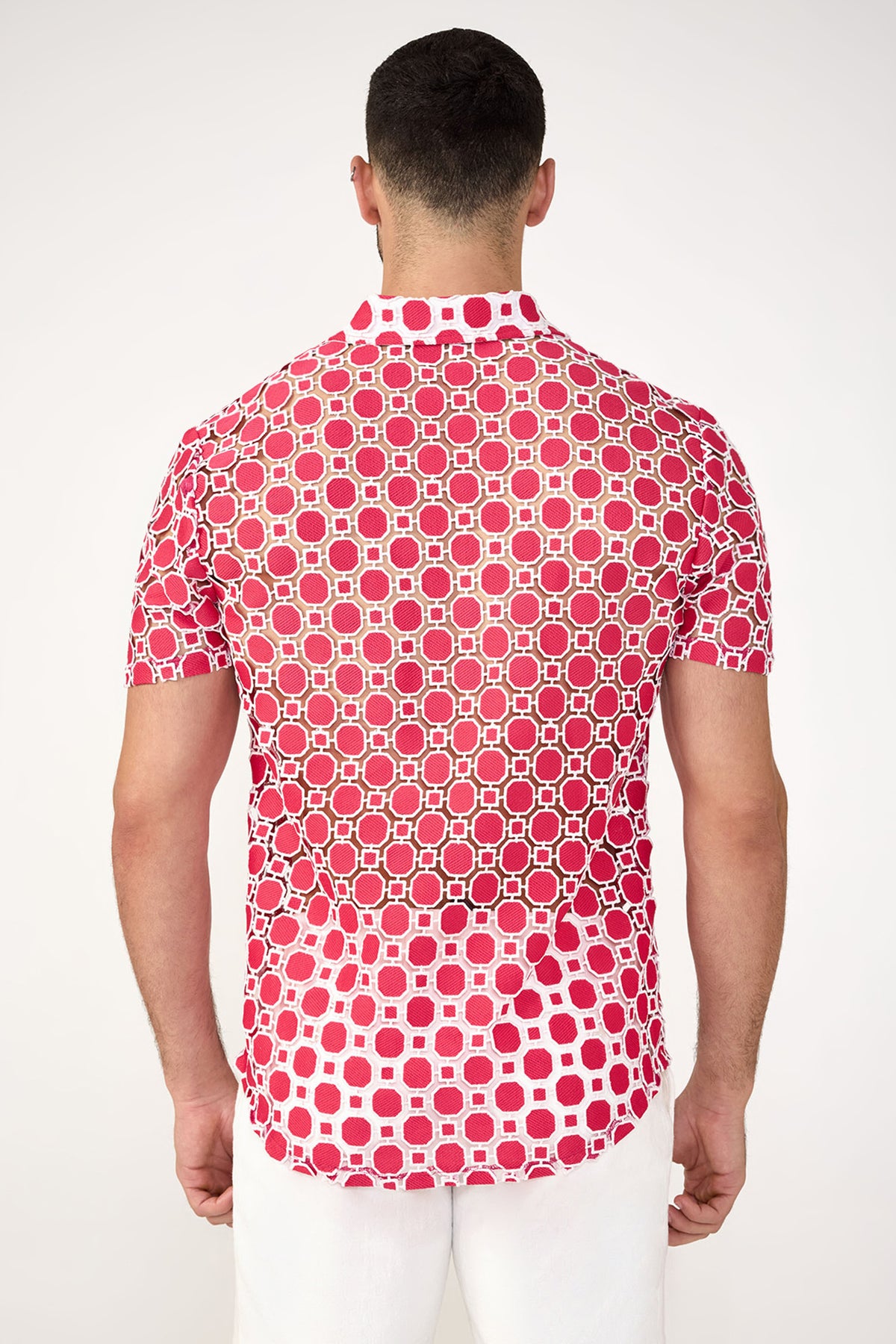 A man stands with his back to the camera, wearing the Long Beach Short-Sleeve Shirt featuring a red and white geometric pattern and white pants against a plain light background.