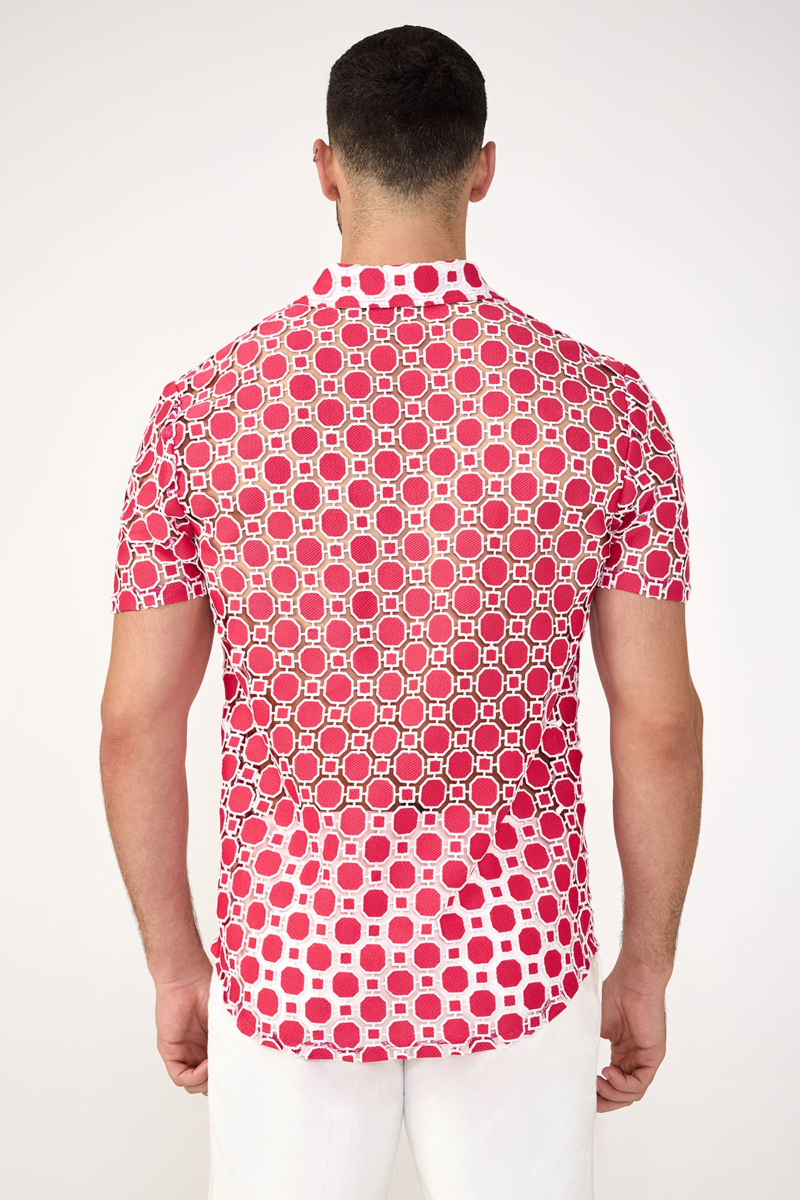 A man stands with his back to the camera, wearing the Long Beach Short-Sleeve Shirt featuring a red and white geometric pattern and white pants against a plain light background.