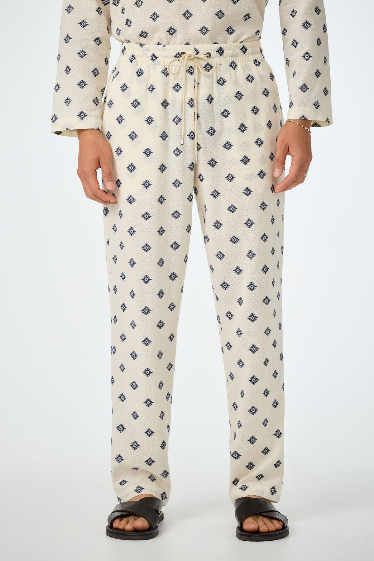 A person wearing Felipe Drawstring Pant in ecru with a blue geometric pattern, matching top, black sandals, and a bracelet, shown from shoulders to feet against a plain background.
