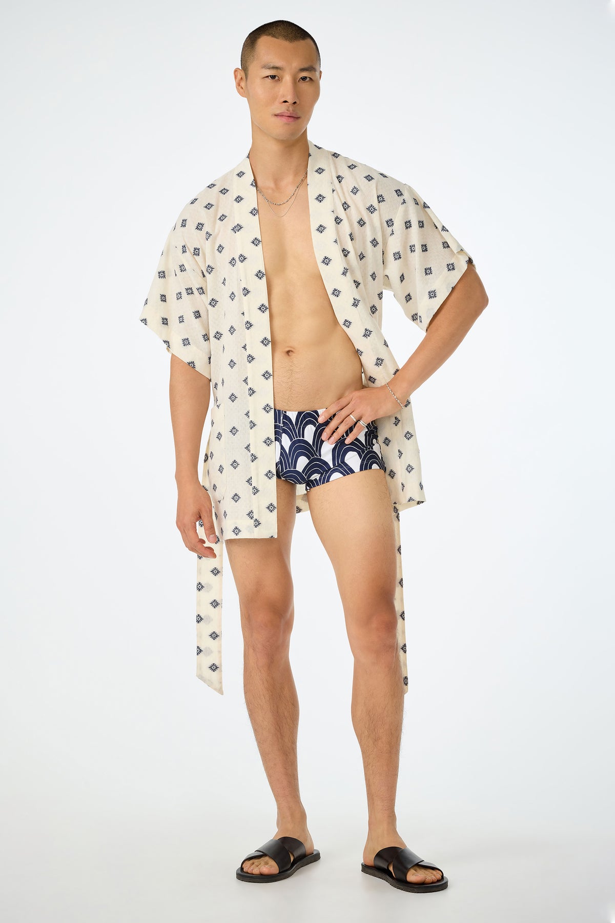 A person stands confidently in an ecru Happi Coat, navy-patterned swim trunks, black sandals, and a thin necklace against a plain white background.