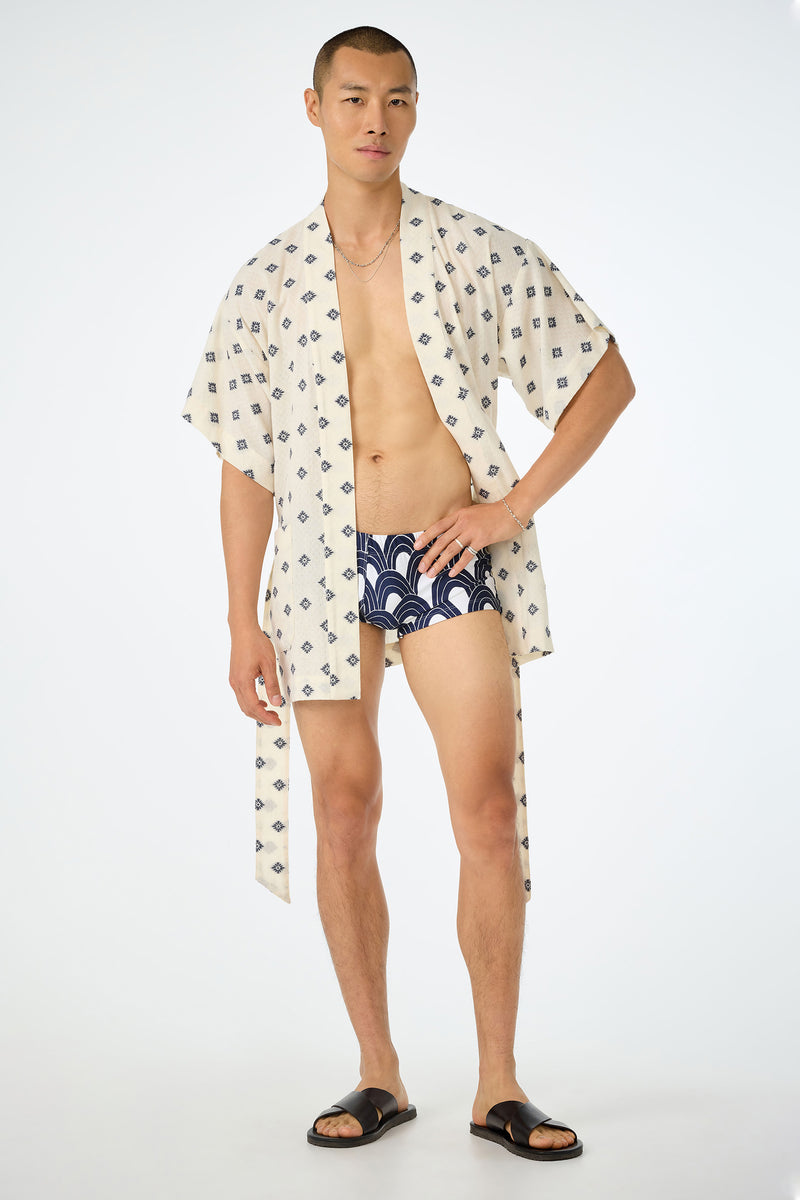 A person stands confidently in an ecru Happi Coat, navy-patterned swim trunks, black sandals, and a thin necklace against a plain white background.