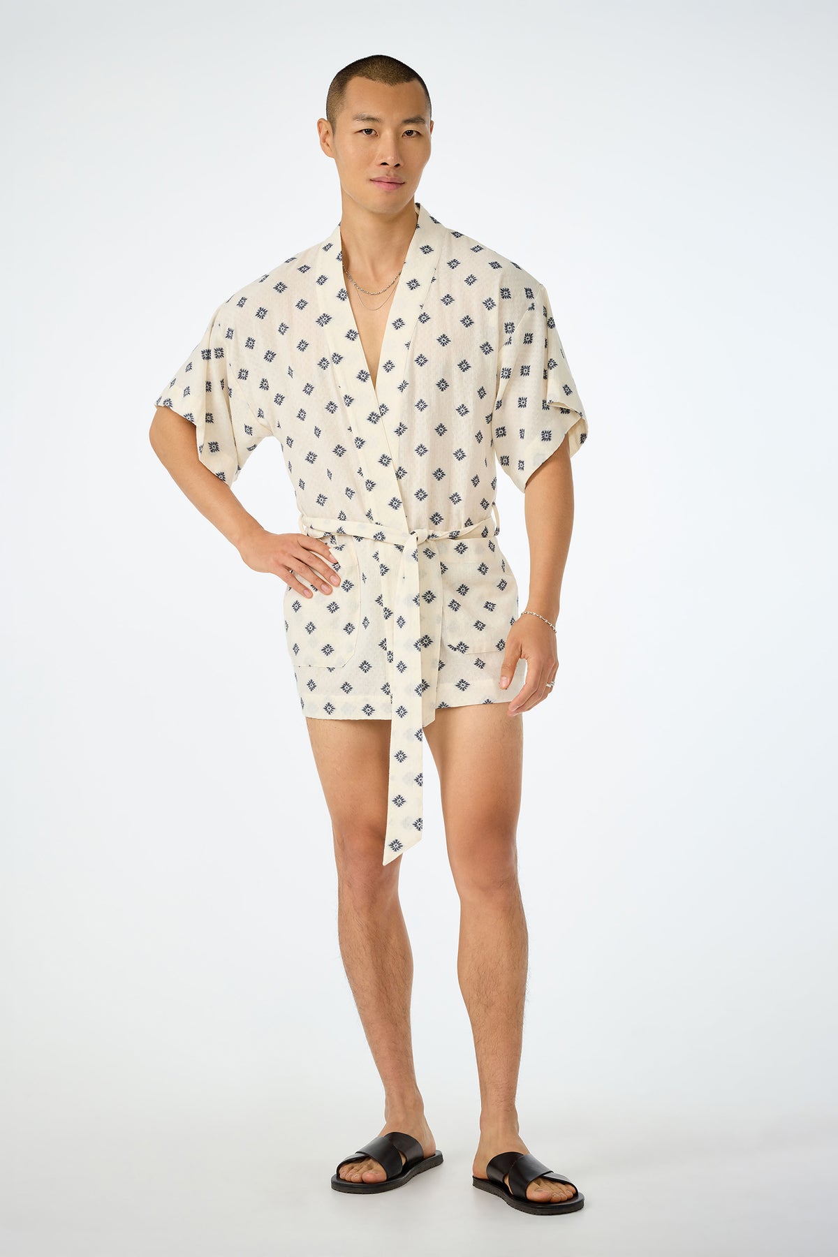 A man stands against a plain background wearing the Happi Coat‚Äîa short, cream-colored romper with a dark geometric pattern, short sleeves, a tie belt‚Äîpaired with black sandals and gold jewelry.