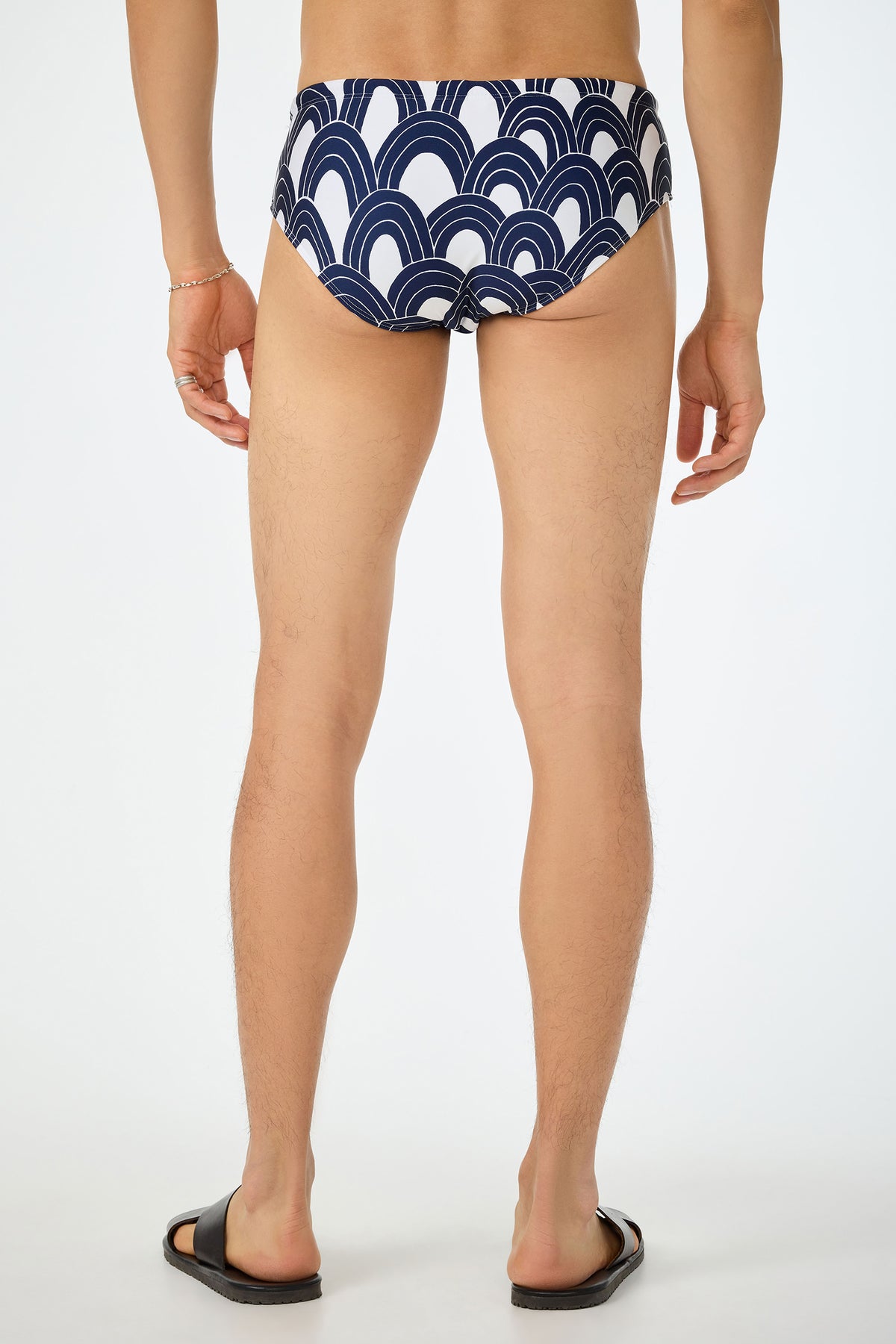 A person stands barefoot in black slide sandals, wearing the Bondi Swim Brief - Arches (white with a navy blue curved wave pattern), shown from the back against a plain white background.
