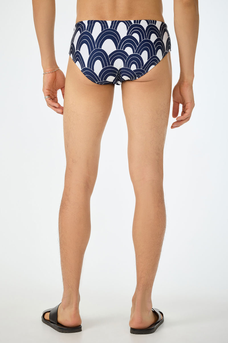 A person stands barefoot in black slide sandals, wearing the Bondi Swim Brief - Arches (white with a navy blue curved wave pattern), shown from the back against a plain white background.