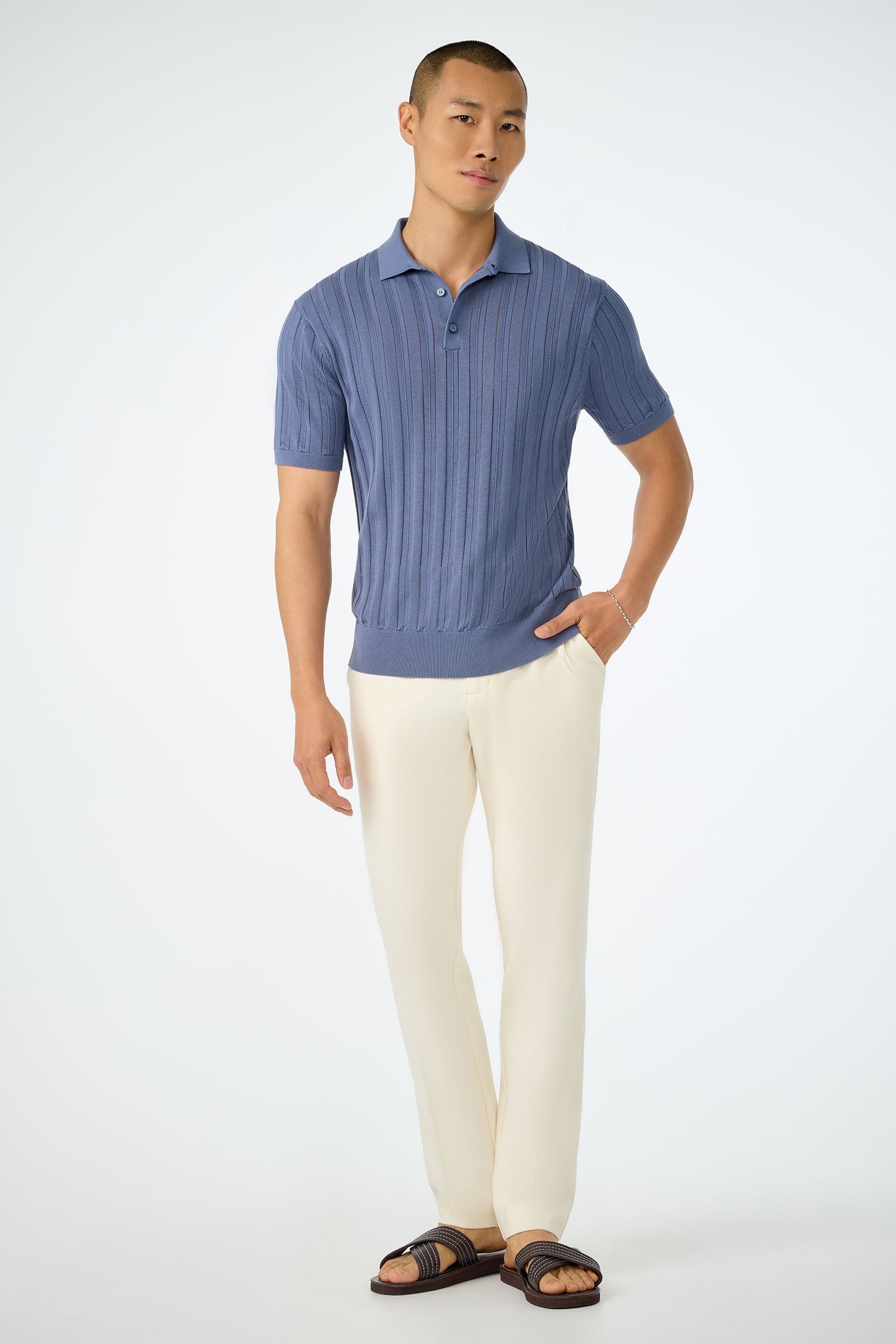 A man with short hair poses against a white background wearing the Kensington Short-Sleeve Polo in blue, cream-colored pants, and black sandals. One hand is in his pocket, and he stands in a relaxed manner.