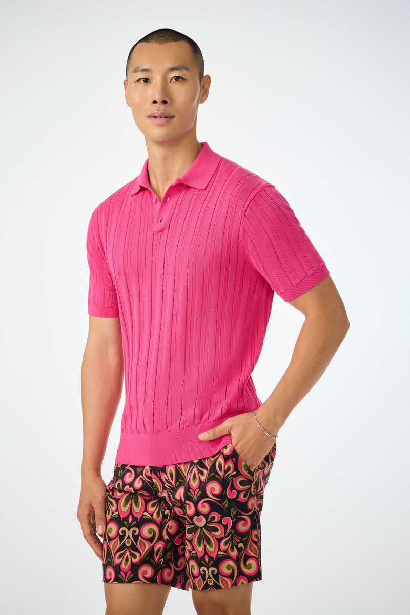 A person stands against a light background wearing the bright pink Kensington Short-Sleeve Polo and colorful patterned shorts with pink, green, and orange swirls, one hand in their pocket.