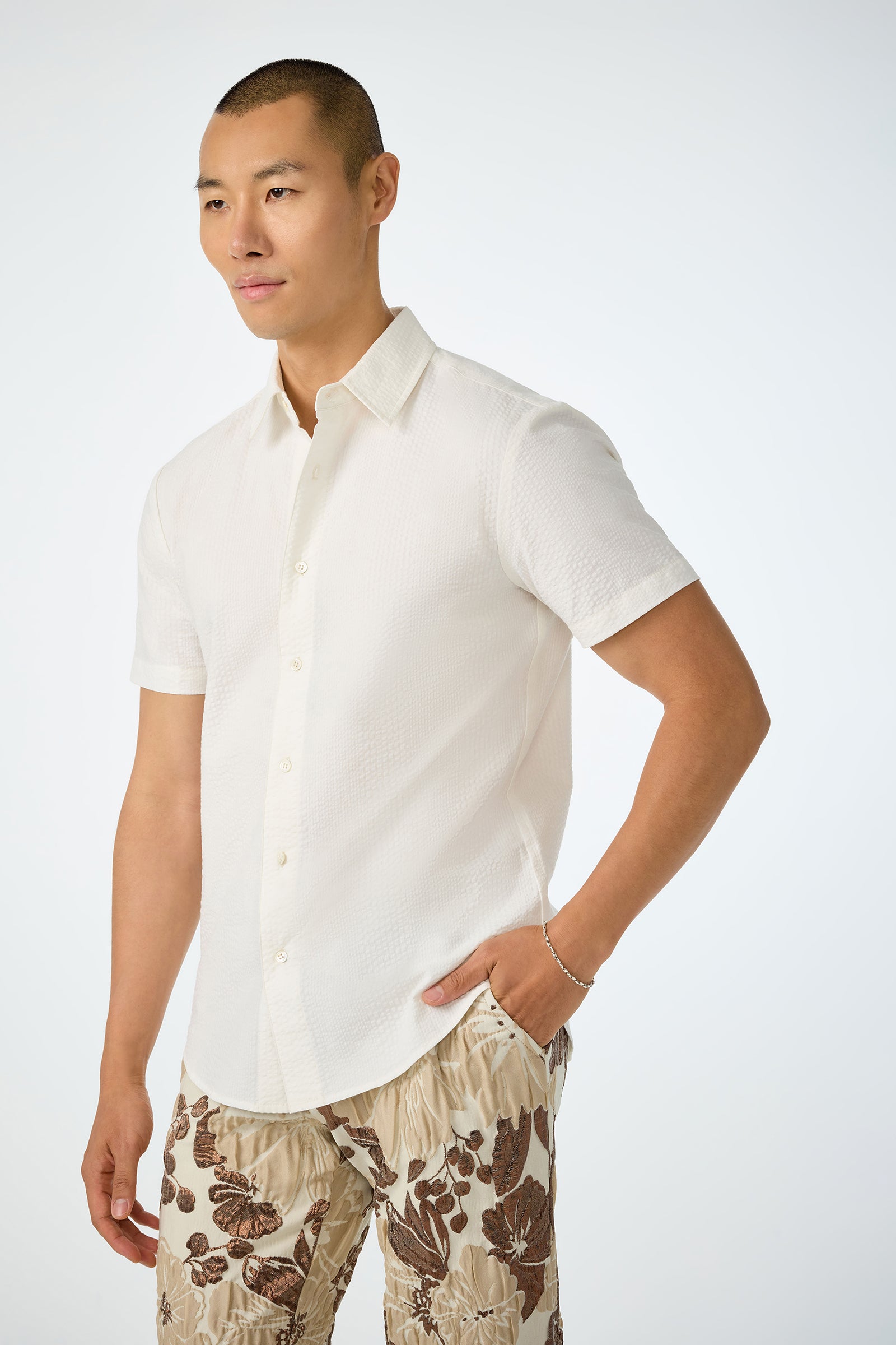 A man with short hair poses against a plain background, wearing the Slim Jim Short-Sleeve Shirt in white and beige floral-patterned shorts, one hand casually in his pocket.