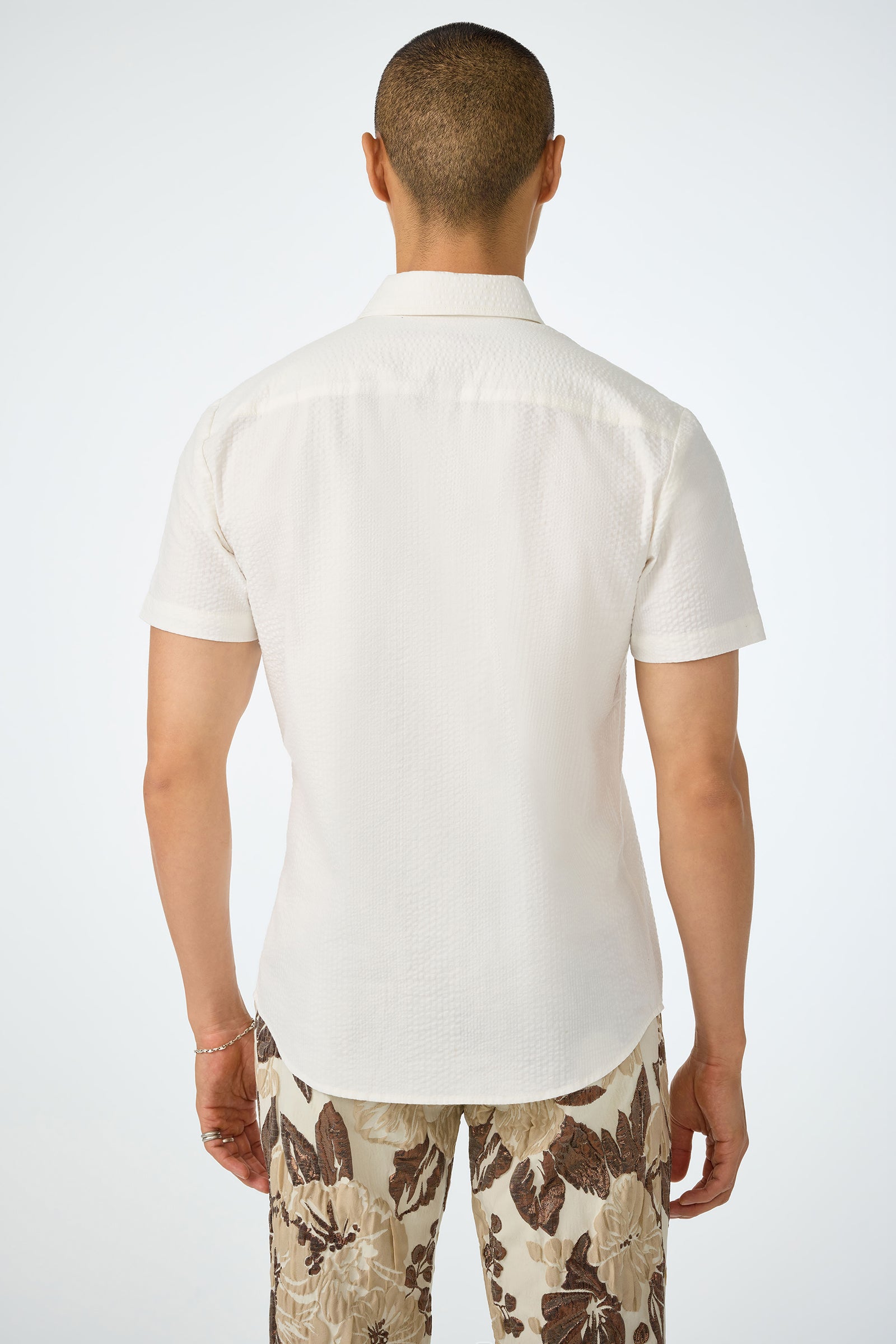 A person stands with their back to the camera, wearing the Slim Jim Short-Sleeve Shirt in white and brown floral-patterned pants against a plain white background.