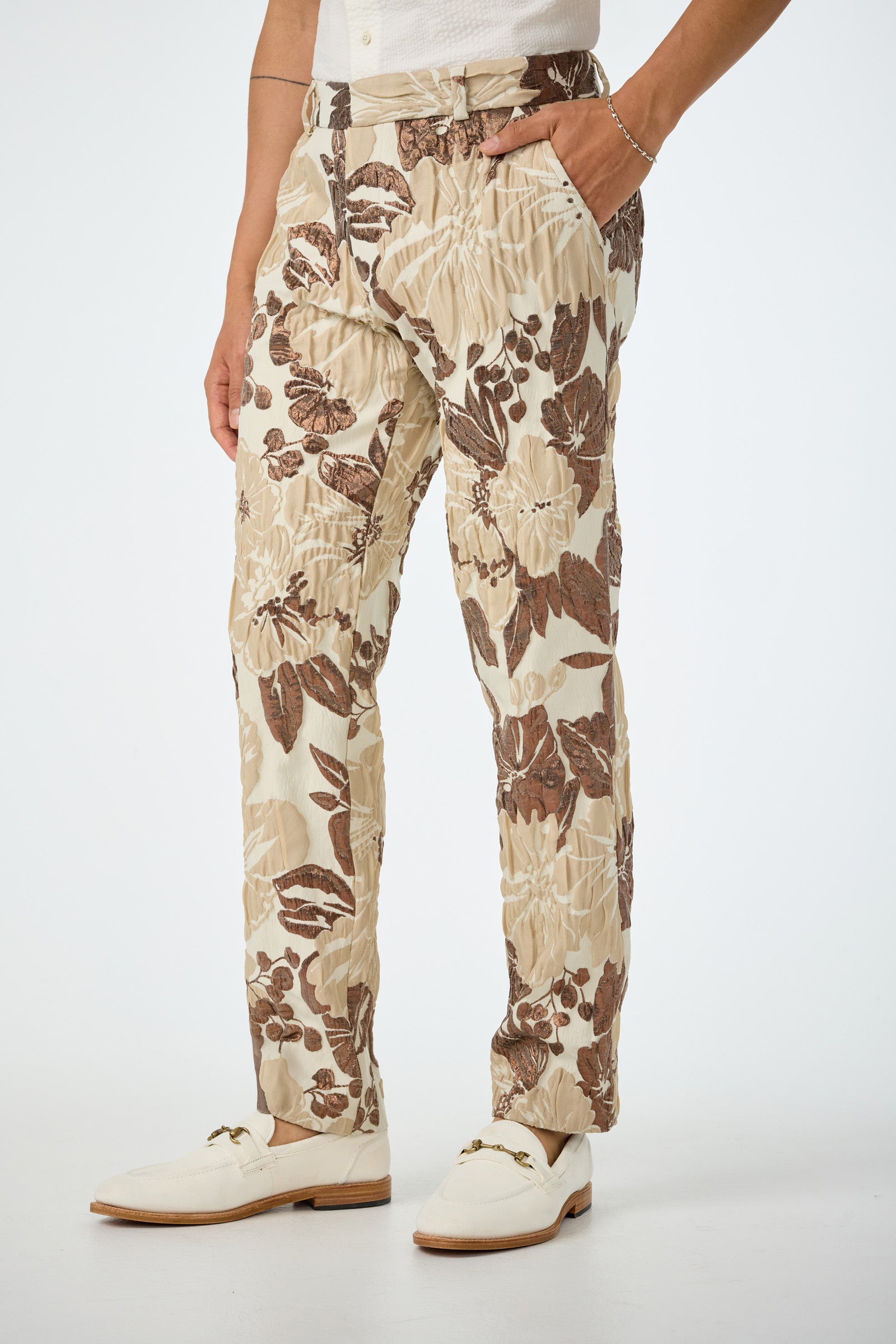 Wearing the Clyde Slim Trouser in Daintree Jacquard—a cream pant with a brown floral pattern—paired with a white shirt, white shoes, and a bracelet, this person stands against a plain white background.