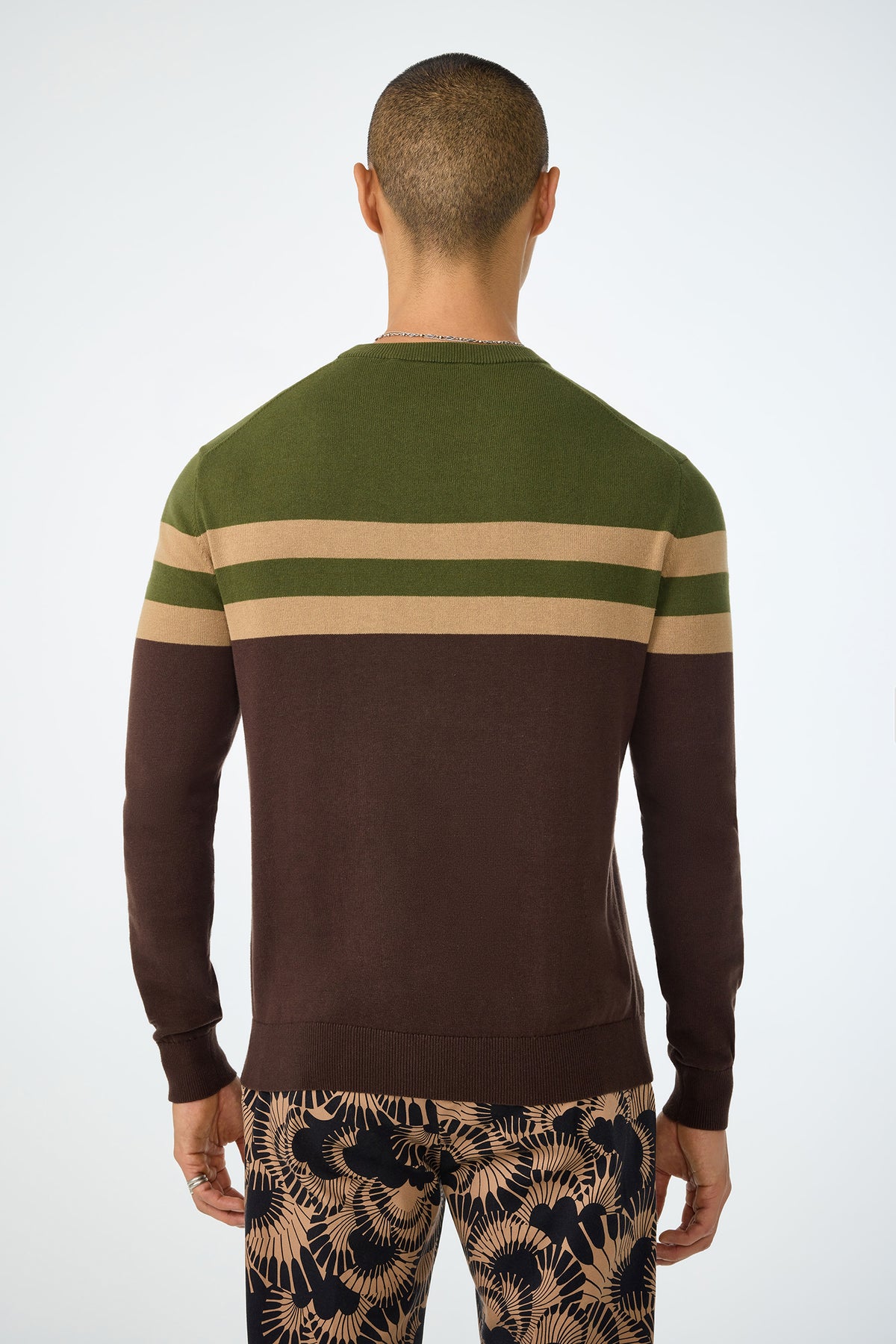 A person with short hair is shown from behind, wearing the Romero Crewneck Sweater in green and brown with beige stripes, and patterned brown leafy-design pants. The background is plain white.