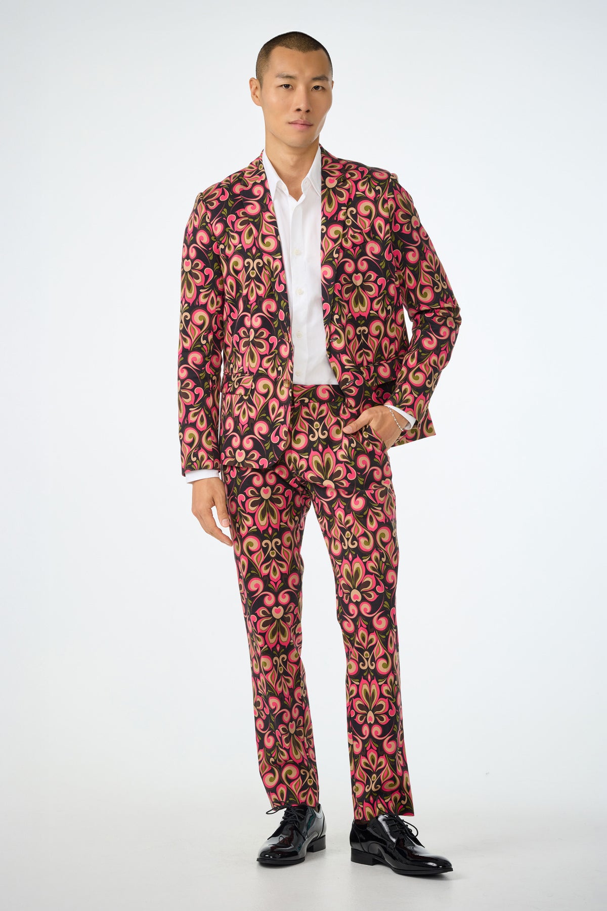 A man stands against a plain background wearing the Clyde Slim Trouser in Sanctuary, featuring a bold, swirling pink, yellow, and black pattern, paired with a white shirt and black dress shoes.
