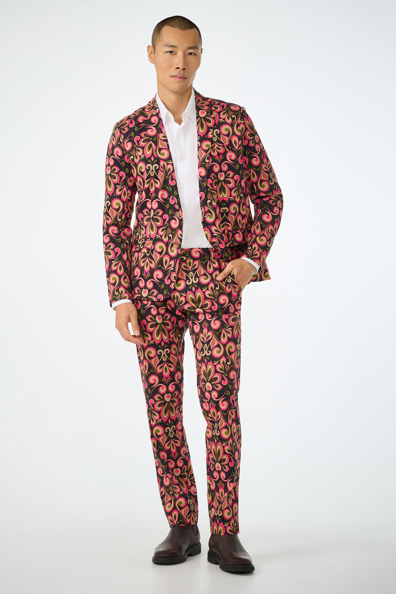 A man wears the Thurston Blazer - Sanctuary, featuring a bold pink, yellow, and black floral pattern, styled with a white shirt and dark shoes, standing against a plain white background.