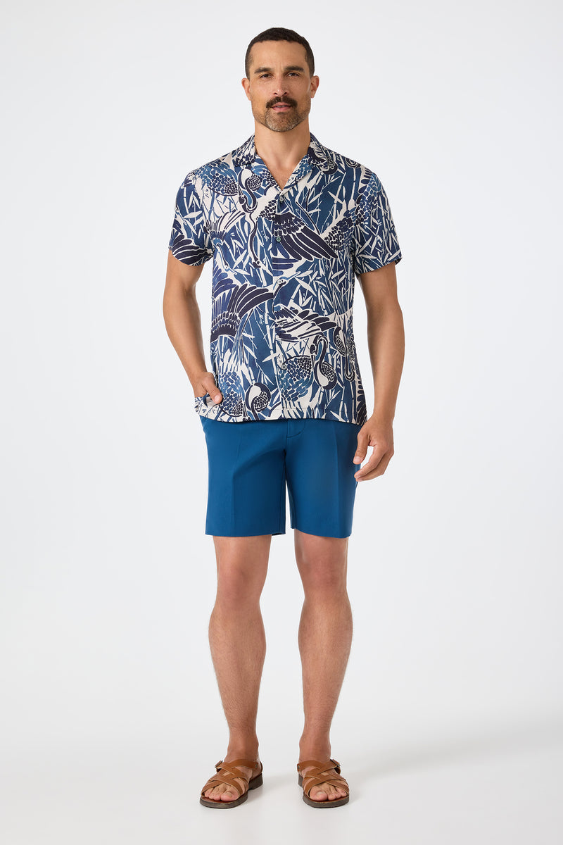 A man with short hair and a mustache stands against a plain background, wearing the Felix Camp Shirt - Tsuru (a blue and white patterned short-sleeve shirt), blue shorts, brown sandals, and has one hand in his pocket.