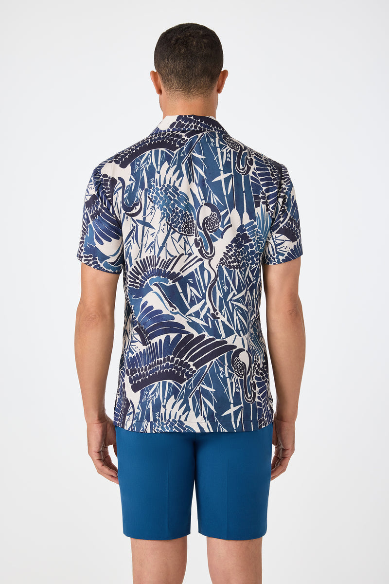 A man stands facing away, wearing the Felix Camp Shirt - Tsuru, a short-sleeve button-up with a blue and white crane pattern, paired with solid blue shorts against a plain white background.