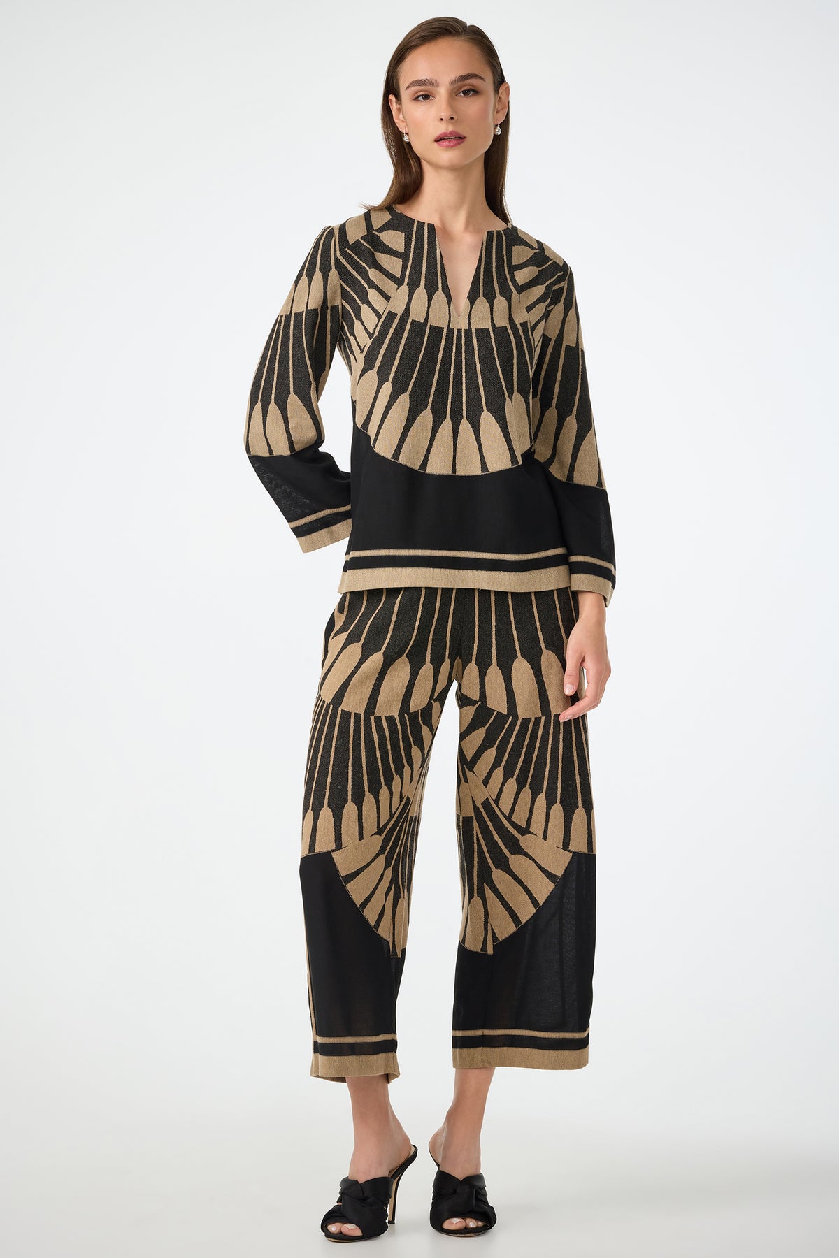 A woman poses against a plain white background in the Estella 2 Cropped Pants - Juniper Fil Coupe, a beige and black geometric set with a long-sleeve top and wide-leg cropped pants, paired with black open-toe heels.