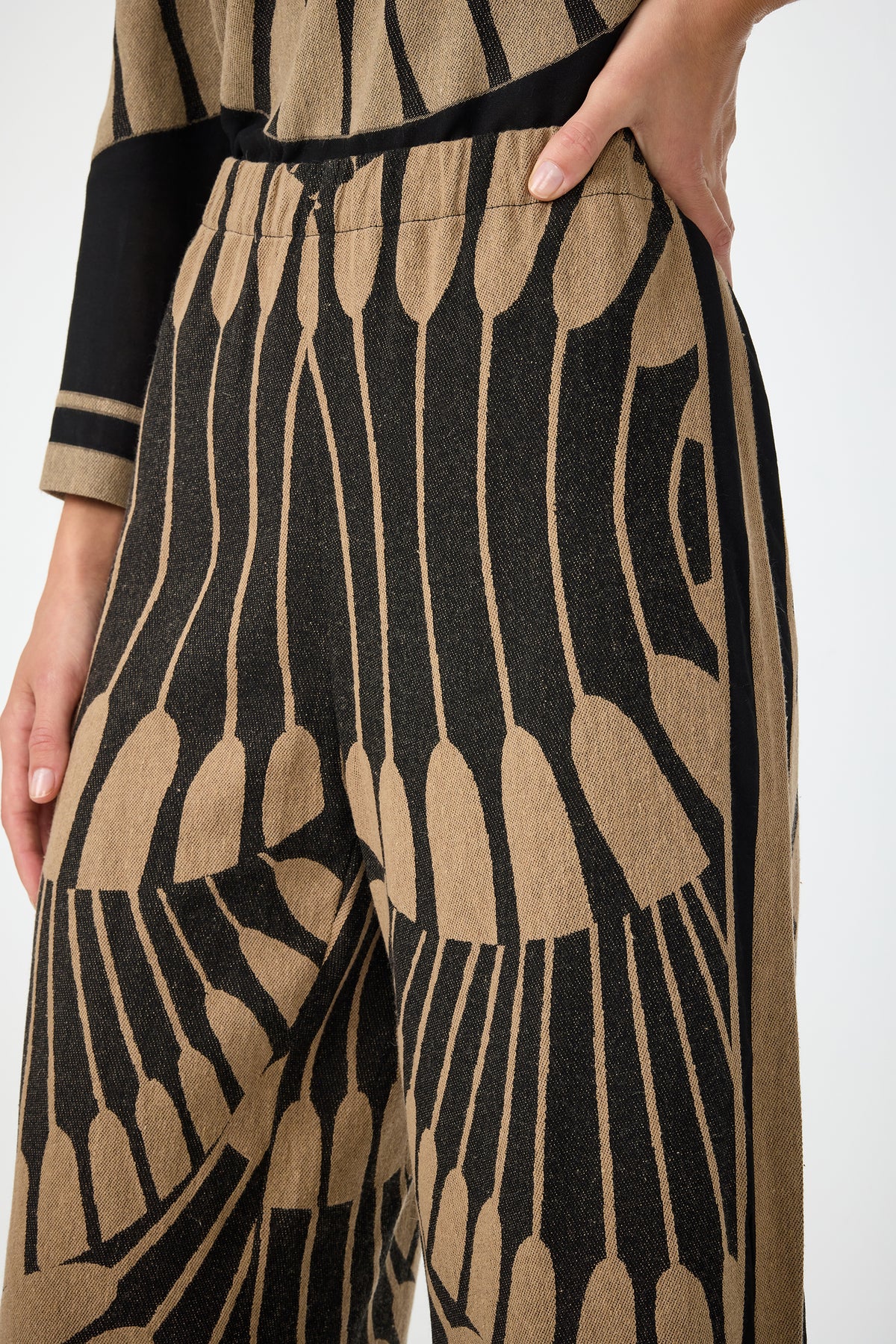 A close-up of a person wearing the Estella 2 Cropped Pants in Juniper Fil Coupe, featuring a bold black and tan abstract pattern. Their hand rests on their hip, and part of a matching top is visible.