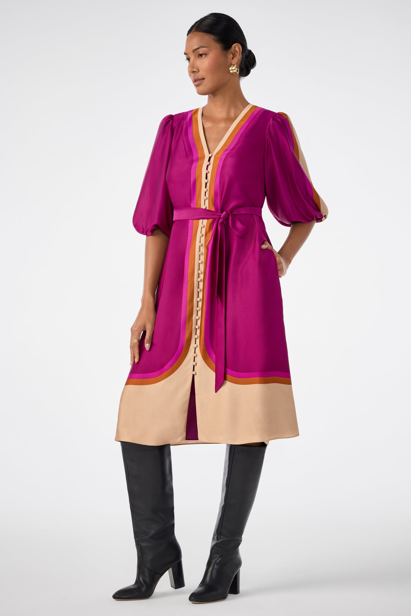 A woman wears the Linden Dress—knee-length, magenta and beige with puffed sleeves, a V-neckline, button details, and a tie belt—paired with black knee-high boots and gold earrings.