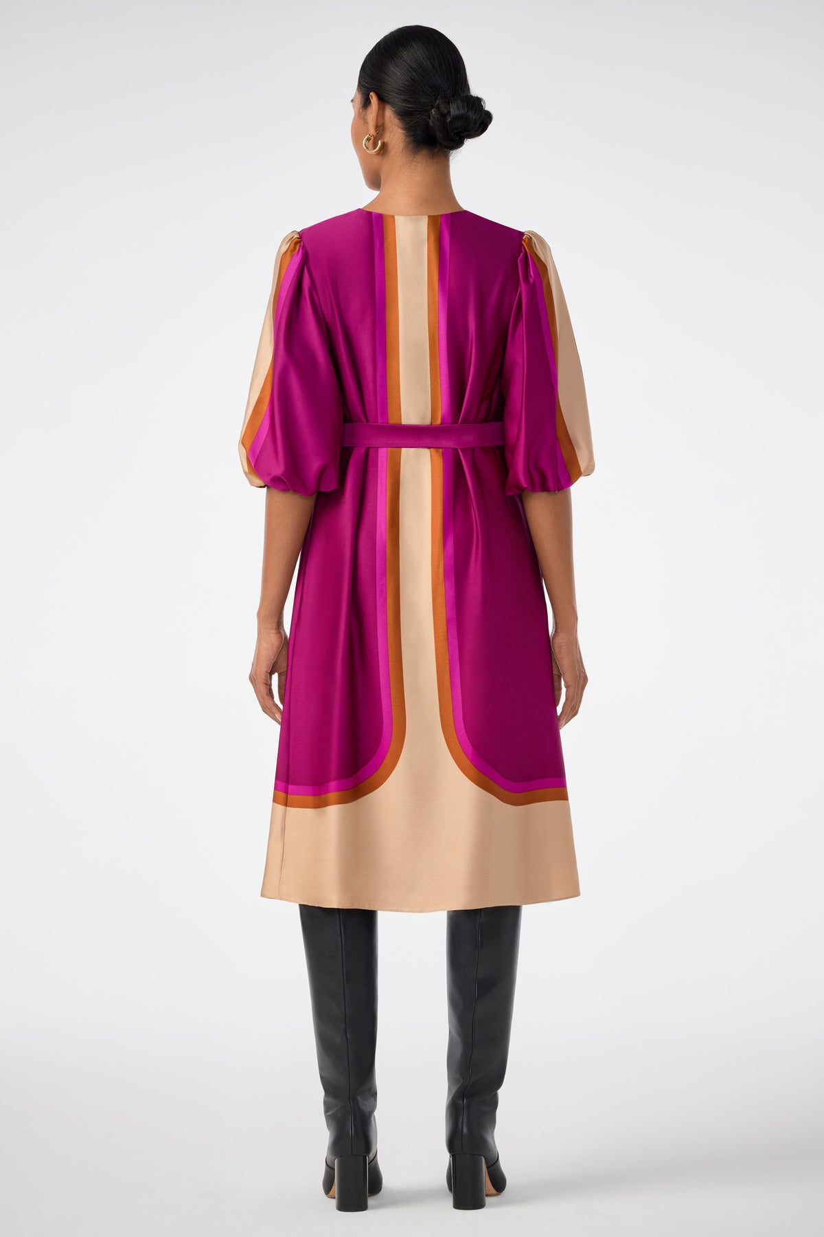 A woman stands facing away, wearing the knee-length Linden Dress with pink, gold, and orange geometric patterns, puffed sleeves, a matching belt, black knee-high boots, and her hair in a low bun.