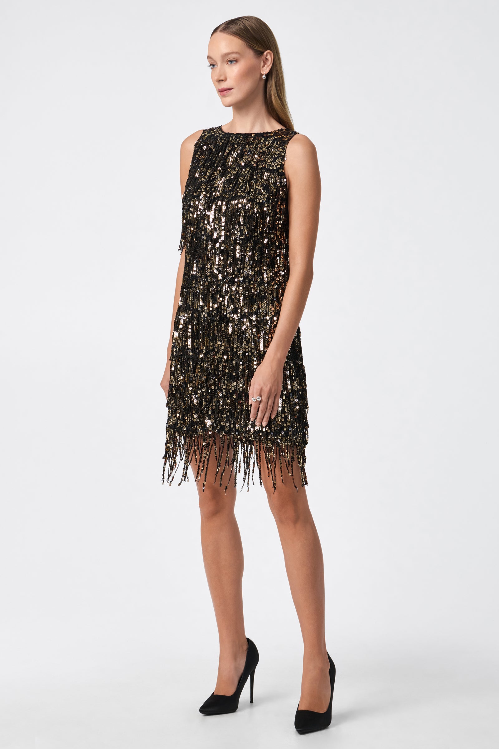 A woman poses against a plain white background wearing the Nini Dress—a sleeveless, knee-length black dress with gold and black sequins, fringe detailing, and paired with black high heels.