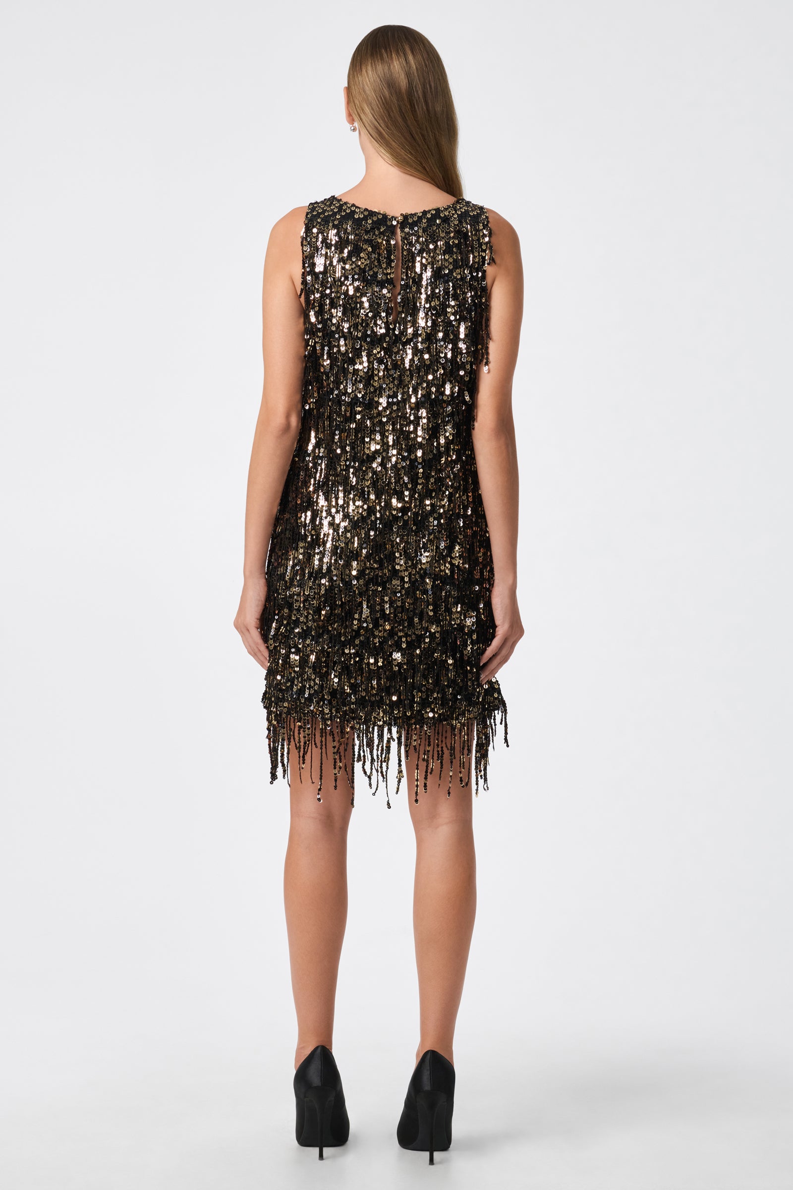 A woman stands with her back to the camera, wearing the Nini Dress—a sleeveless black dress adorned with gold sequins and fringe—paired with black heels. Her straight hair is loose, set against a plain white background.