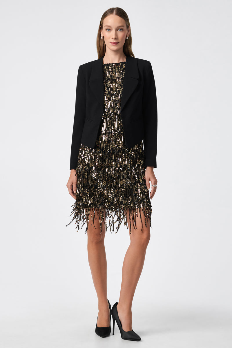 A woman faces forward in a black Ren Cropped Blazer over a short gold and black sequin fringe dress. She has straight brown hair, wears black high heels, and stands against a plain white background.