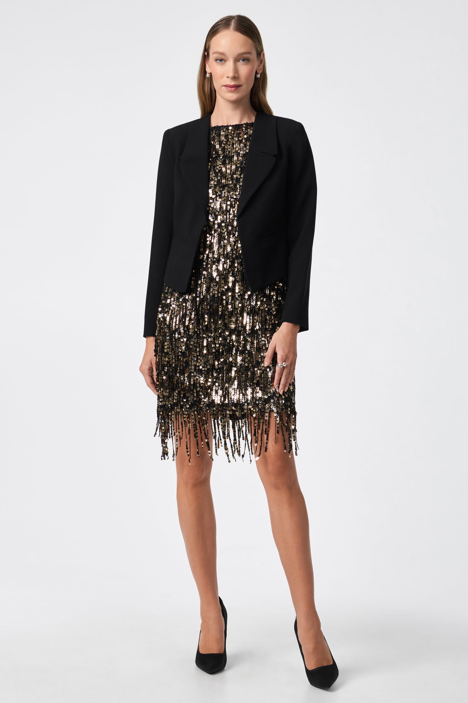 A woman in the Nini Dress—a shiny, sequined gold and black fringe dress—styled with a black blazer and high heels, stands against a plain white background.
