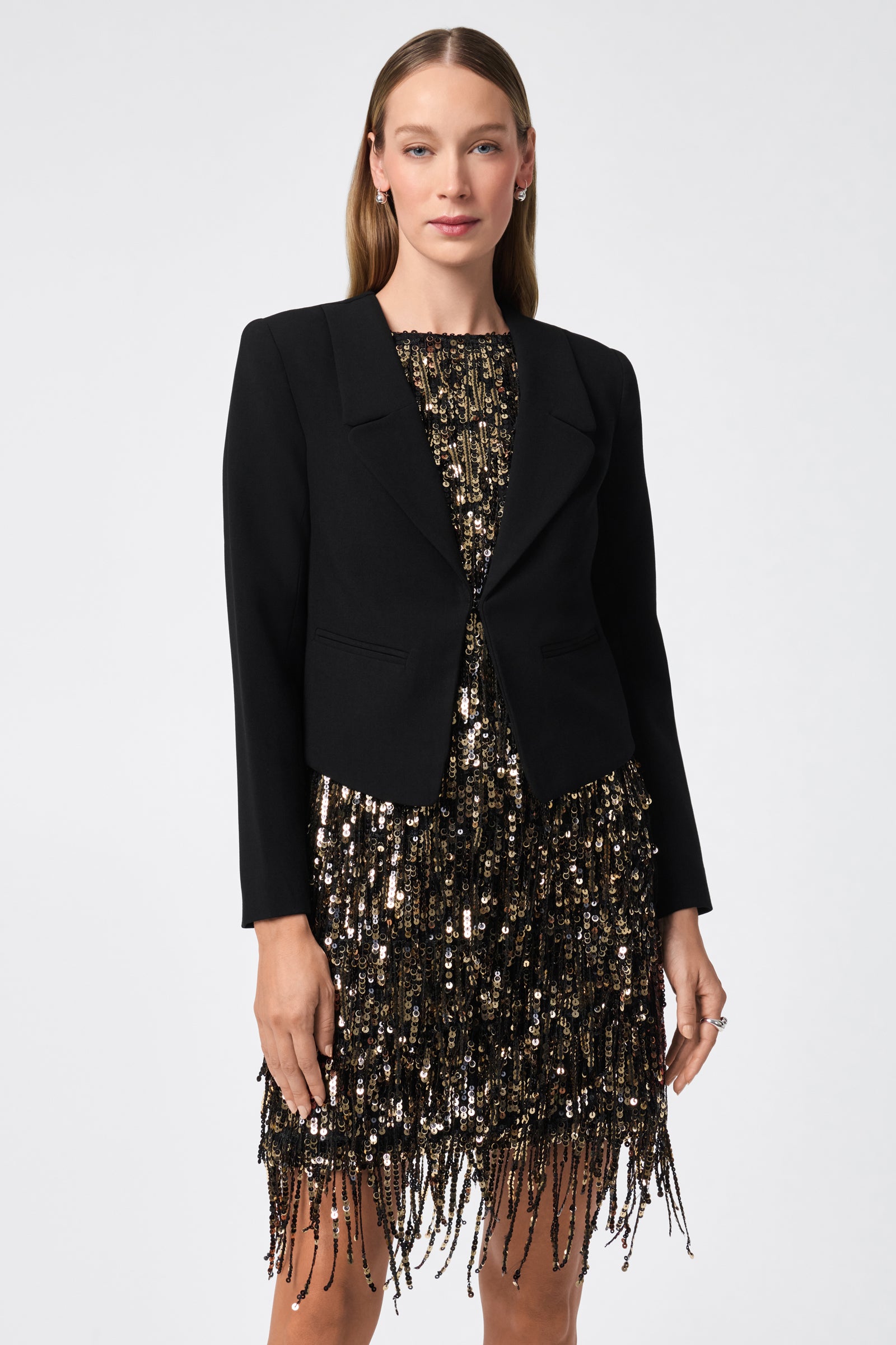 A woman faces forward, wearing the Ren Cropped Long Sleeve Blazer Jacket over a knee-length gold and black sequined dress with fringe. She has straight, light brown hair and a neutral expression against a plain background.