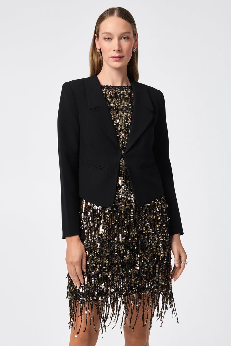 A woman faces forward, wearing the Ren Cropped Long Sleeve Blazer Jacket over a knee-length gold and black sequined dress with fringe. She has straight, light brown hair and a neutral expression against a plain background.