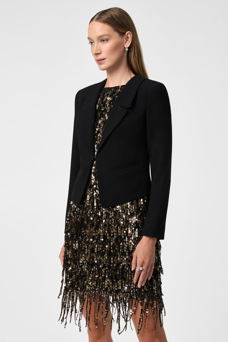 A woman with straight light brown hair poses against a plain white background, wearing the Ren Cropped Long Sleeve Blazer Jacket over a black and gold sequined fringe dress.