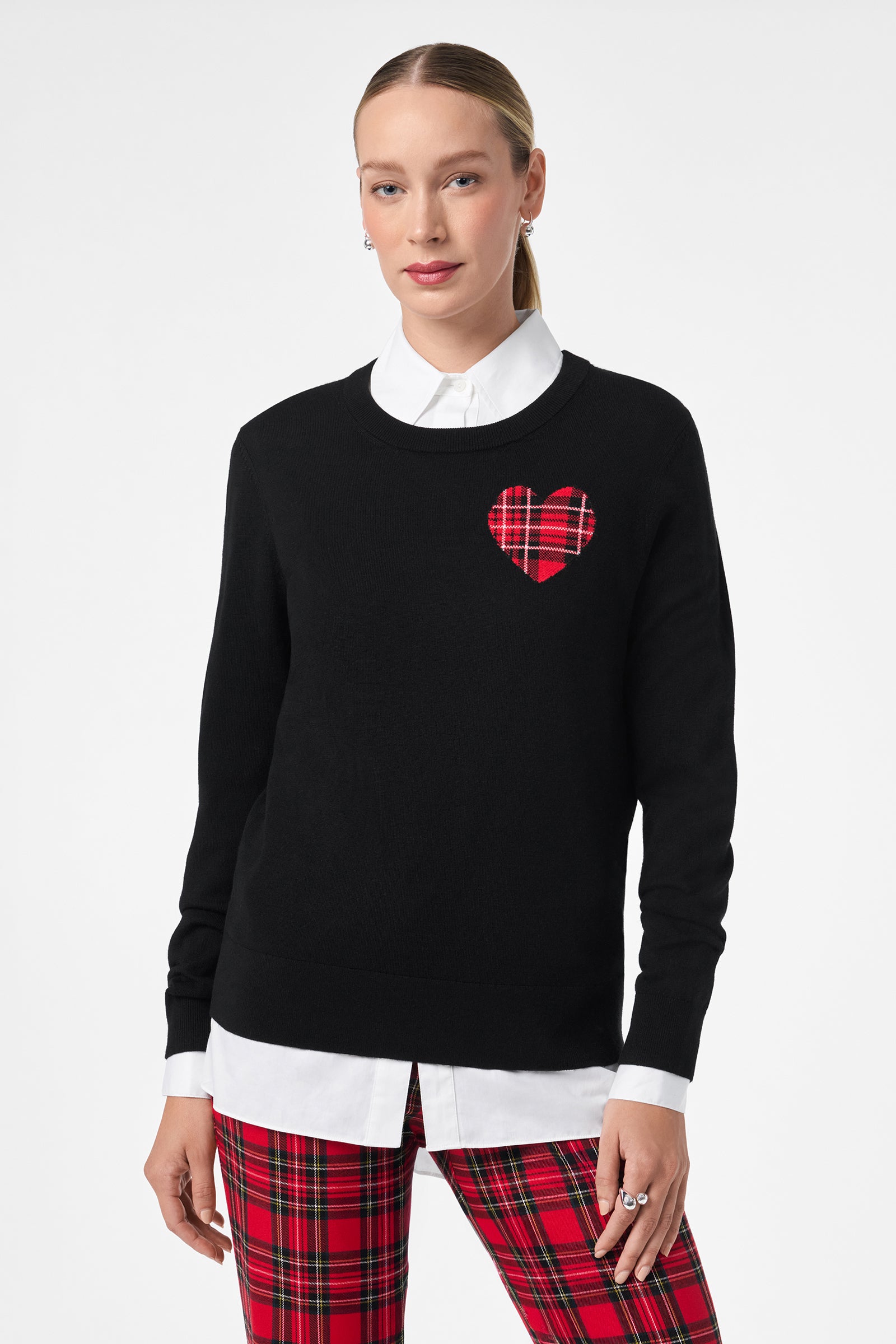 A woman wearing the Plaid Heart Sweater—a black sweater with a red plaid heart patch—layered over a white collared shirt and paired with red plaid pants, stands against a plain white background.