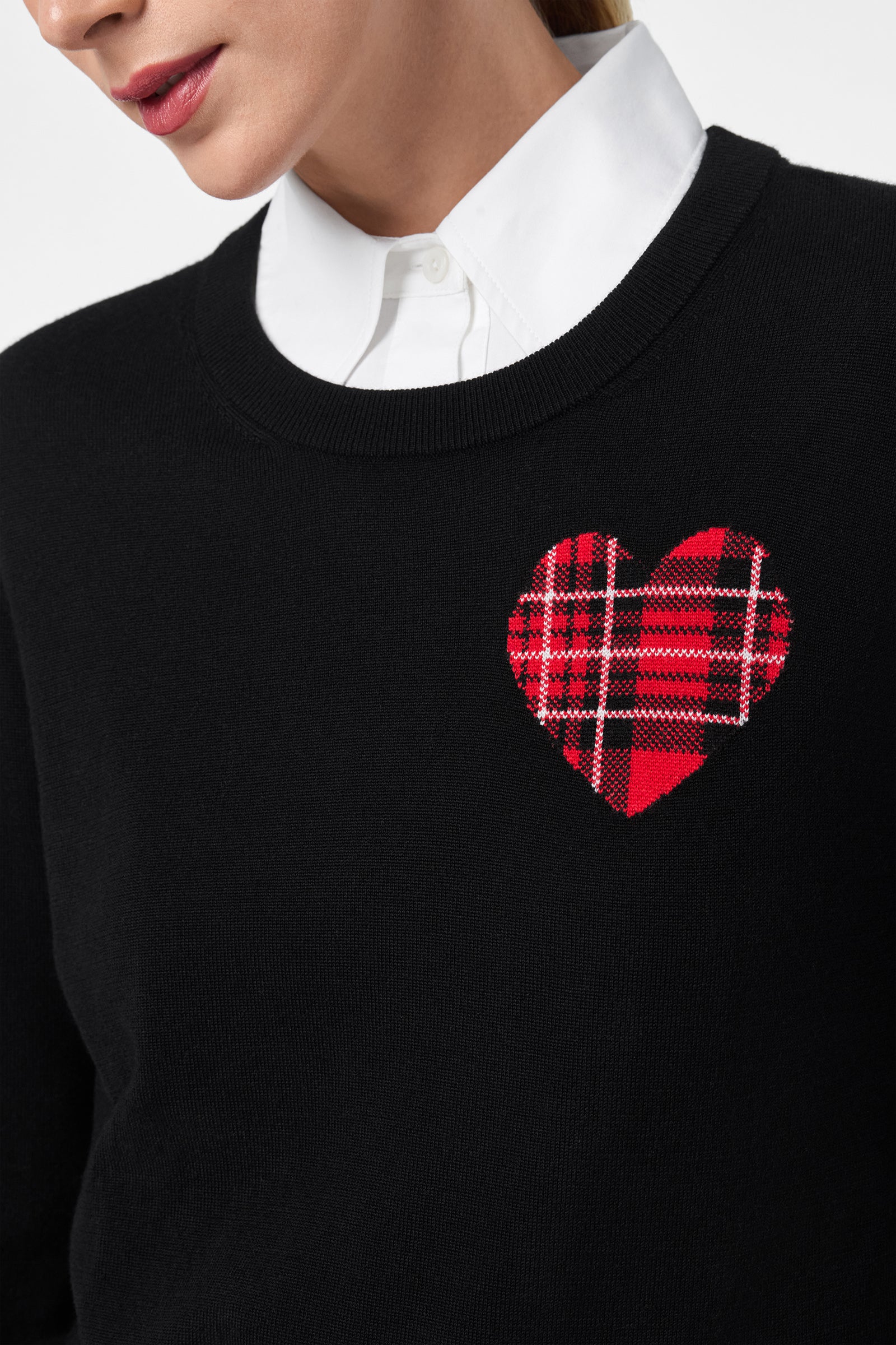 A woman models the Plaid Heart Sweater, which features a red plaid heart patch on the chest and is styled over a white collared shirt. Only her lower face and upper torso are shown.