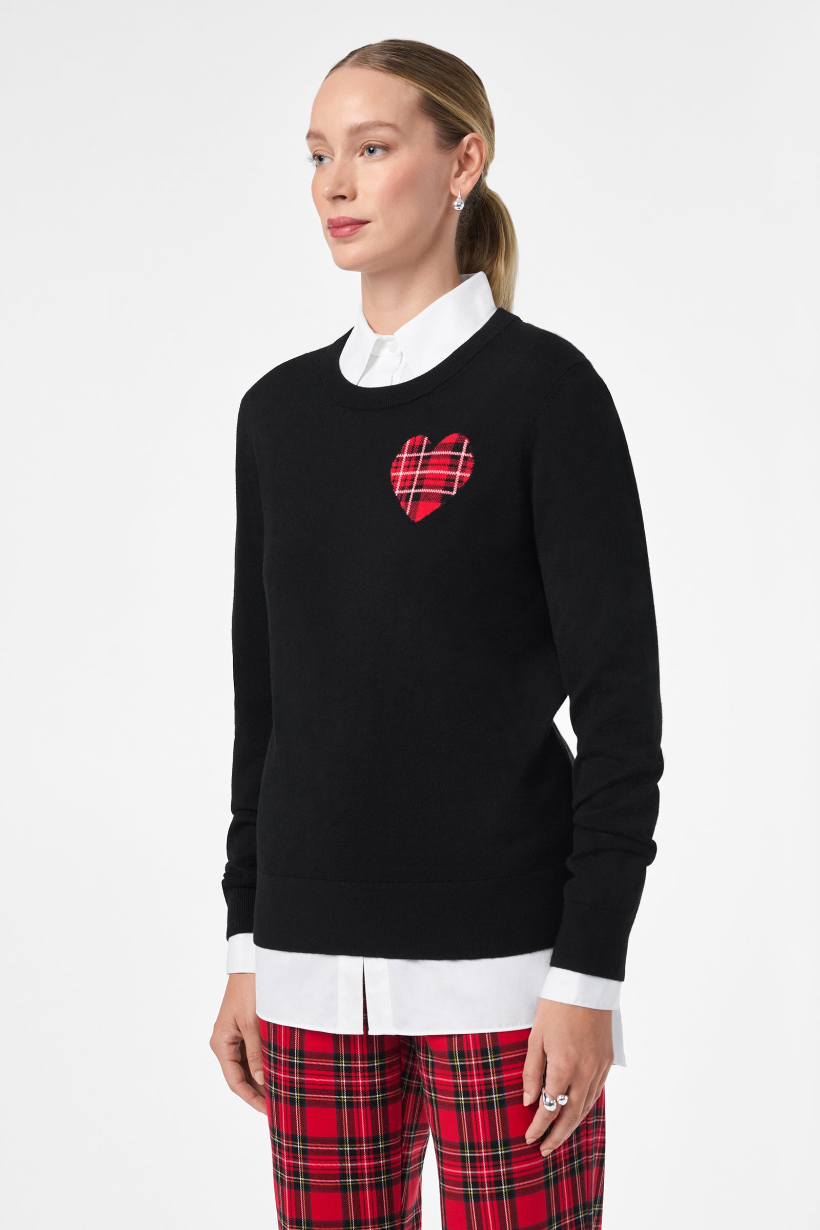 A woman wears the Plaid Heart Sweater—black with a red plaid heart patch—over a white collared shirt and red plaid pants, standing against a plain white background.