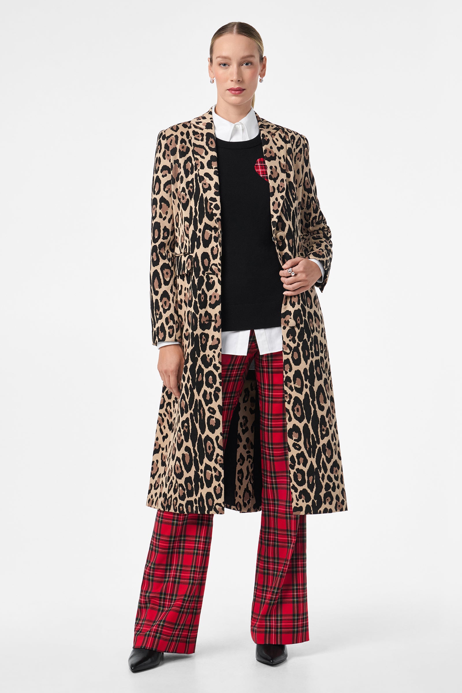 A woman faces forward against a plain white background, wearing the Plaid Heart Sweater beneath a long leopard-print coat, paired with a white collared shirt, red plaid pants, and black pointed shoes.