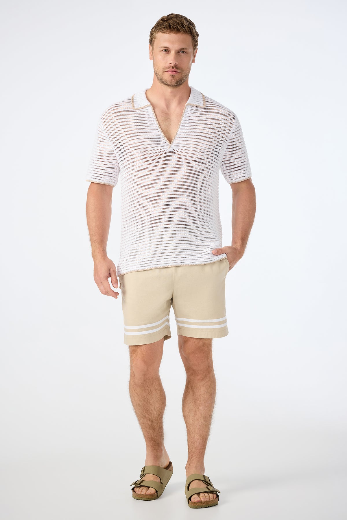 A man with light brown hair and a trimmed beard stands facing forward in a Triston Short-Sleeve Crochet Polo, beige striped shorts, and olive green sandals against a plain white background.