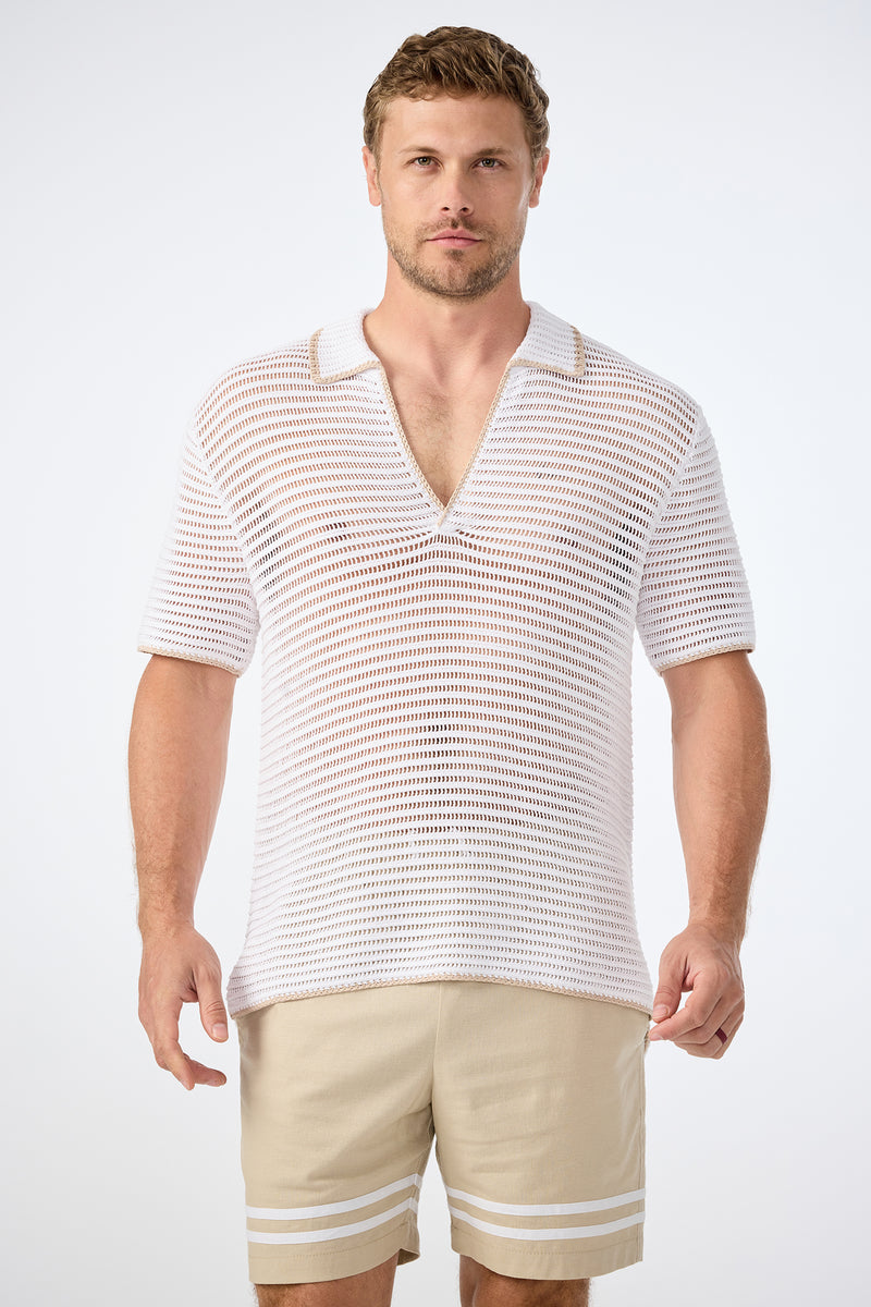 A man faces forward in a plain, light setting, wearing the Triston Short-Sleeve Crochet Polo‚Äö√Ñ√Æwhite, collared, V-neck and loosely knit‚Äö√Ñ√Æpaired with light beige shorts featuring white stripes near the hem.