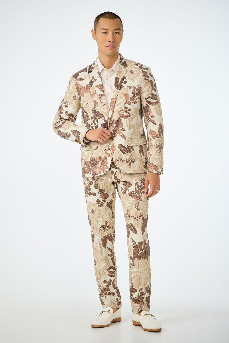 A man stands against a plain background wearing the Thurston Blazer - Daintree Jacquard, featuring a bold brown floral pattern over beige, paired with a white dress shirt and shoes. He closes one jacket button and looks confidently at the camera.
