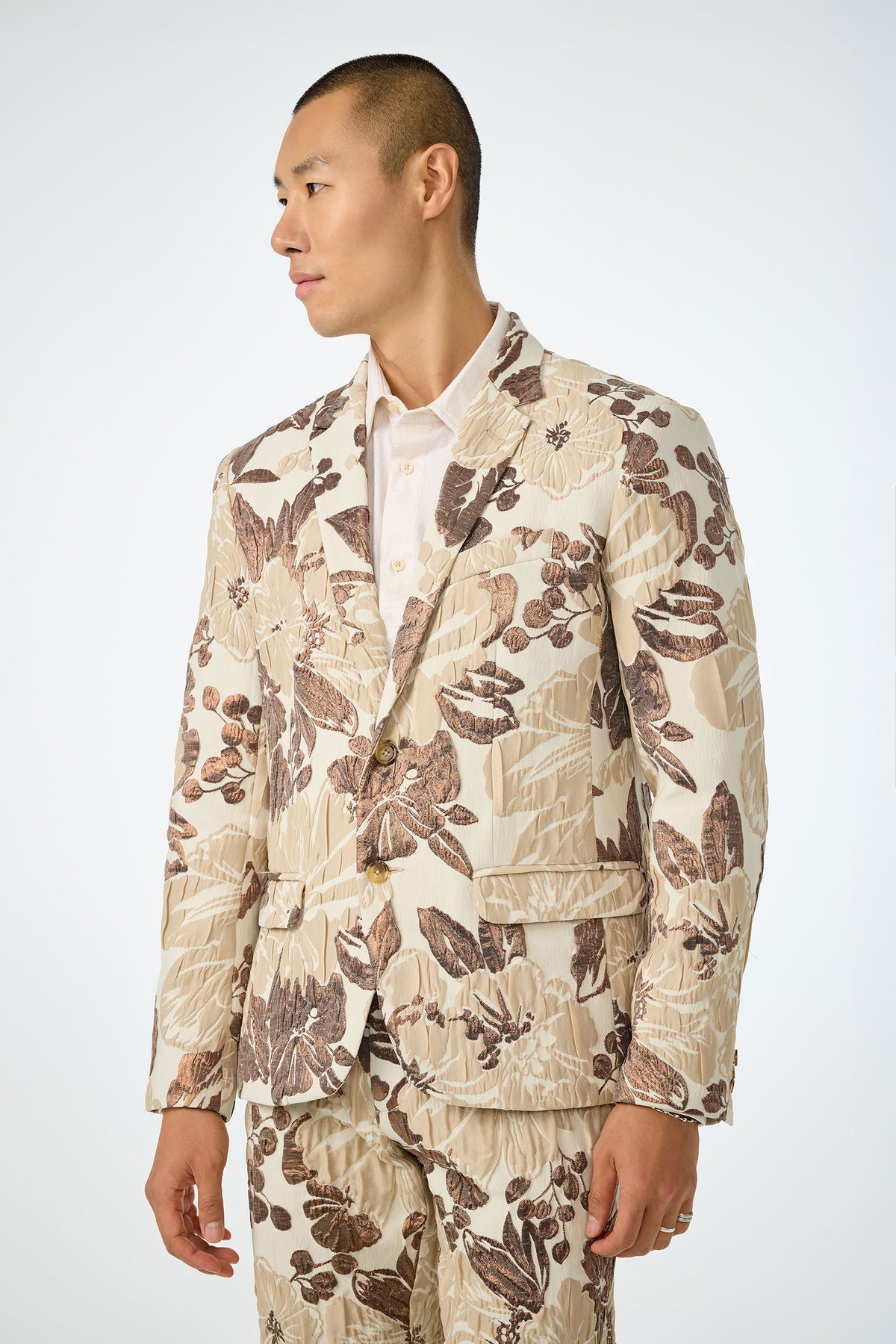 A man with short hair wears the Thurston Blazer - Daintree Jacquard, a cream suit with a bold brown and beige floral pattern, over a light shirt, standing against a plain background and looking to his left.