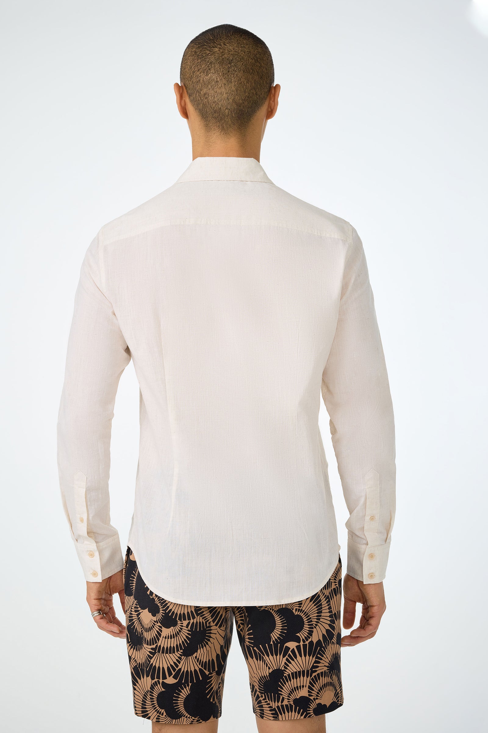 A man with a shaved head is shown from the back, wearing the Aaron Slim Fit Long-Sleeve Shirt in a light color and dark shorts with a beige leaf pattern, standing against a plain white background.