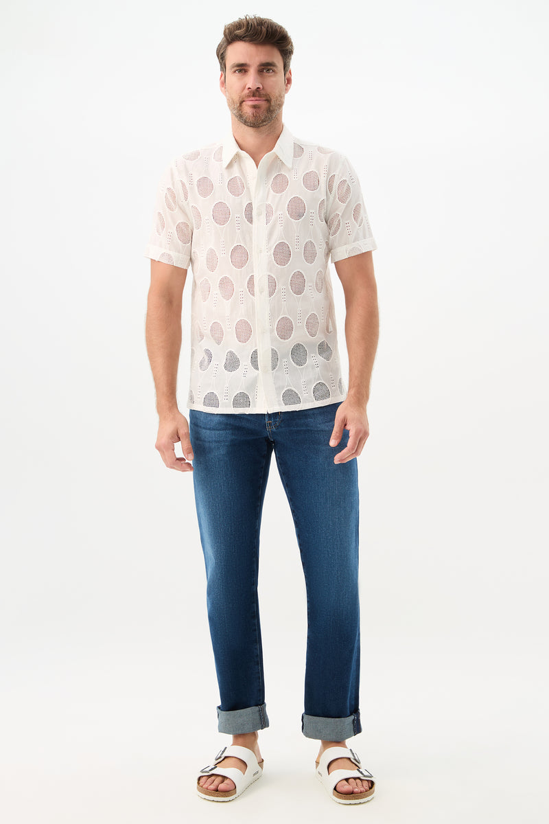 A man stands facing forward against a plain white background, wearing the Branson Shirt - Tennis Eyelet (a white, short-sleeve, see-through shirt with circular patterns), blue cuffed jeans, and white slide sandals.