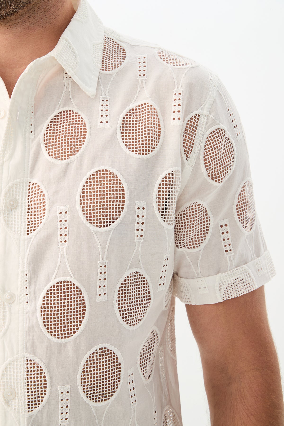 A close-up of a person wearing the Branson Shirt - Tennis Eyelet, a white short-sleeve button-up with mesh cutouts featuring repeating tennis racket and ball patterns, subtly revealing skin underneath.