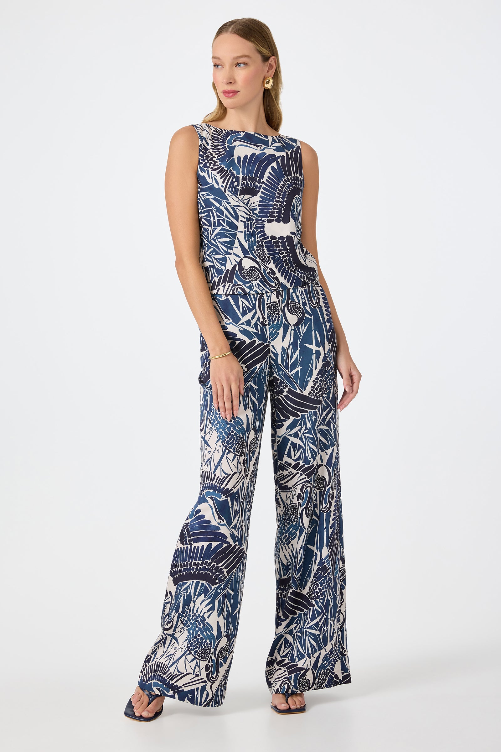 A woman with long blonde hair stands against a white background, wearing the Tavia Wide-Leg Pant - Tsuru featuring a bold navy and white bird and foliage print, paired with sandals.