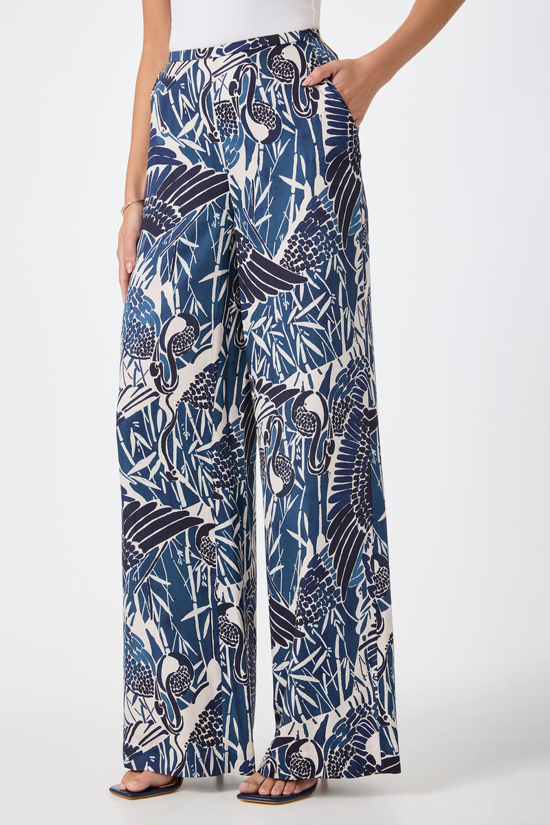 A person wearing the Tavia Wide-Leg Pant - Tsuru, featuring a bold blue, white, and black abstract nature pattern, paired with black sandals and one hand in their pocket.