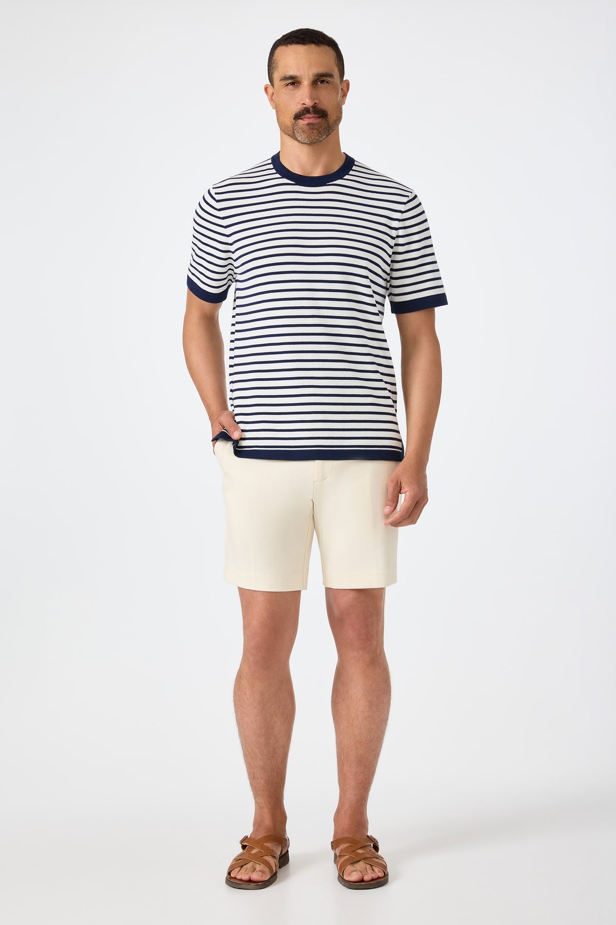 A man with short hair and a mustache wears the Lawrence Short in Ecru with a navy and white striped short-sleeve shirt and brown sandals, standing against a plain background with one hand in his pocket.