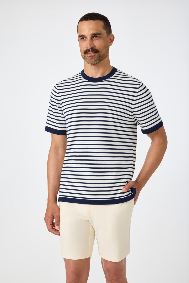 A man with short dark hair and a mustache wears the Giovanni Stripe Short-Sleeve Pullover in indigo, paired with cream shorts, standing with one hand in his pocket against a plain light background.