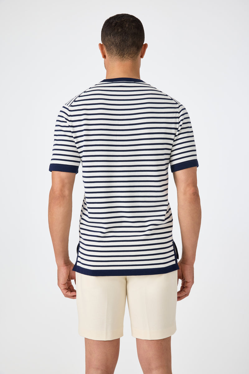 A man faces away from the camera, wearing the Giovanni Stripe Short-Sleeve Pullover‚Äîwhite with navy horizontal stripes and trim‚Äîpaired with light shorts. The background is plain and light.
